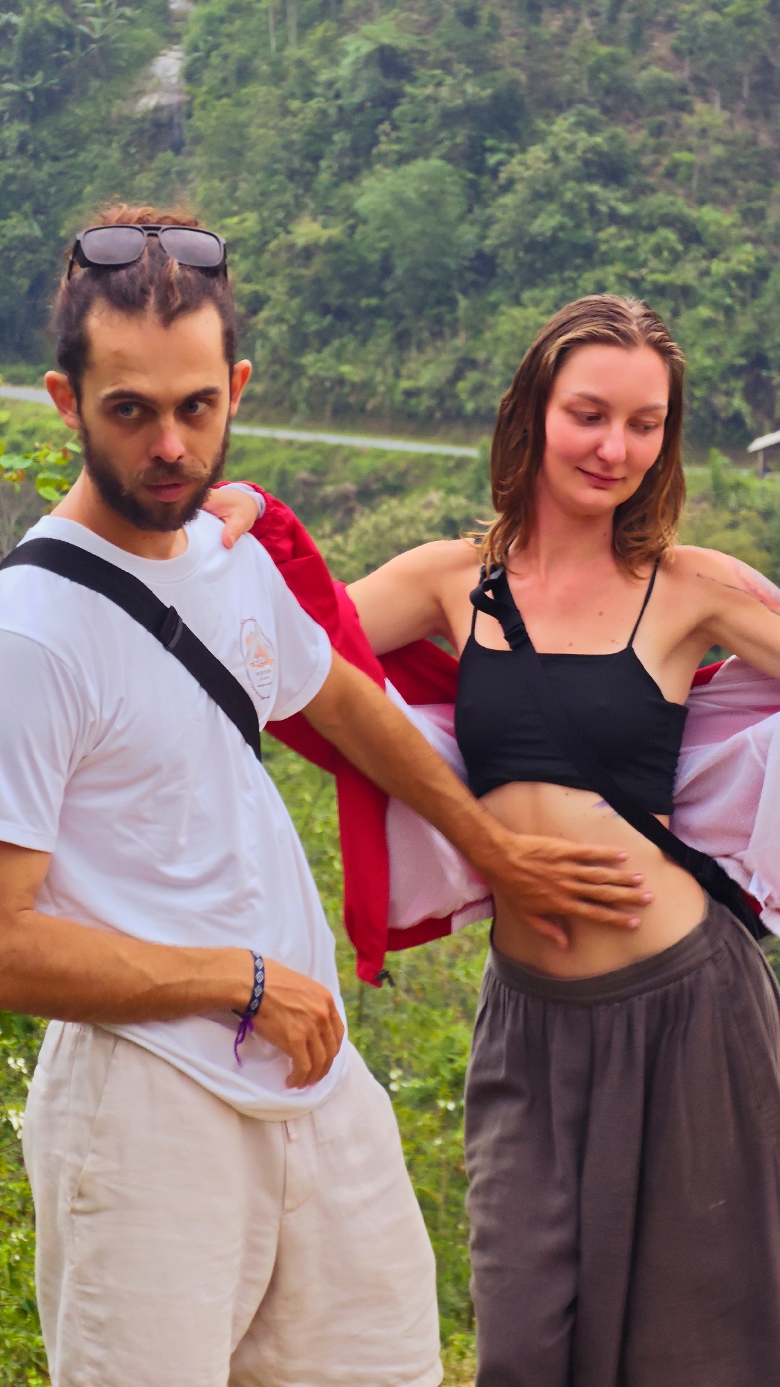 This outdoor portrait features a man and a woman in a lush, mountainous landscape. The man, on the left, has dark hair tied back with sunglasses resting on top, a beard, and a mustache. He wears a white t-shirt with an indistinct graphic on the chest, a black cross-body bag, and light-colored shorts. A dark blue and white woven bracelet is visible on his right wrist. His gaze is direct and intense, facing the viewer.
The woman, on the right, has brown hair and a soft smile as she looks downwards. She wears a black crop top and dark grey, wide-leg pants. A red jacket is draped over her shoulders, held open. The man's right hand is placed on the woman's bare stomach, while her left hand is also near her abdomen.
They appear to be posing together in a natural setting. The background is dominated by dense green foliage, indicating a forest or jungle-covered hillside. A narrow, light-colored road or path winds through the mid-ground. The natural light suggests it is daytime. The overall environment points to a rural or wilderness area, specifically Nậm Dịch Commune, Vietnam, characterized by its verdant mountains and pathways. No clearly legible text is visible in the image.