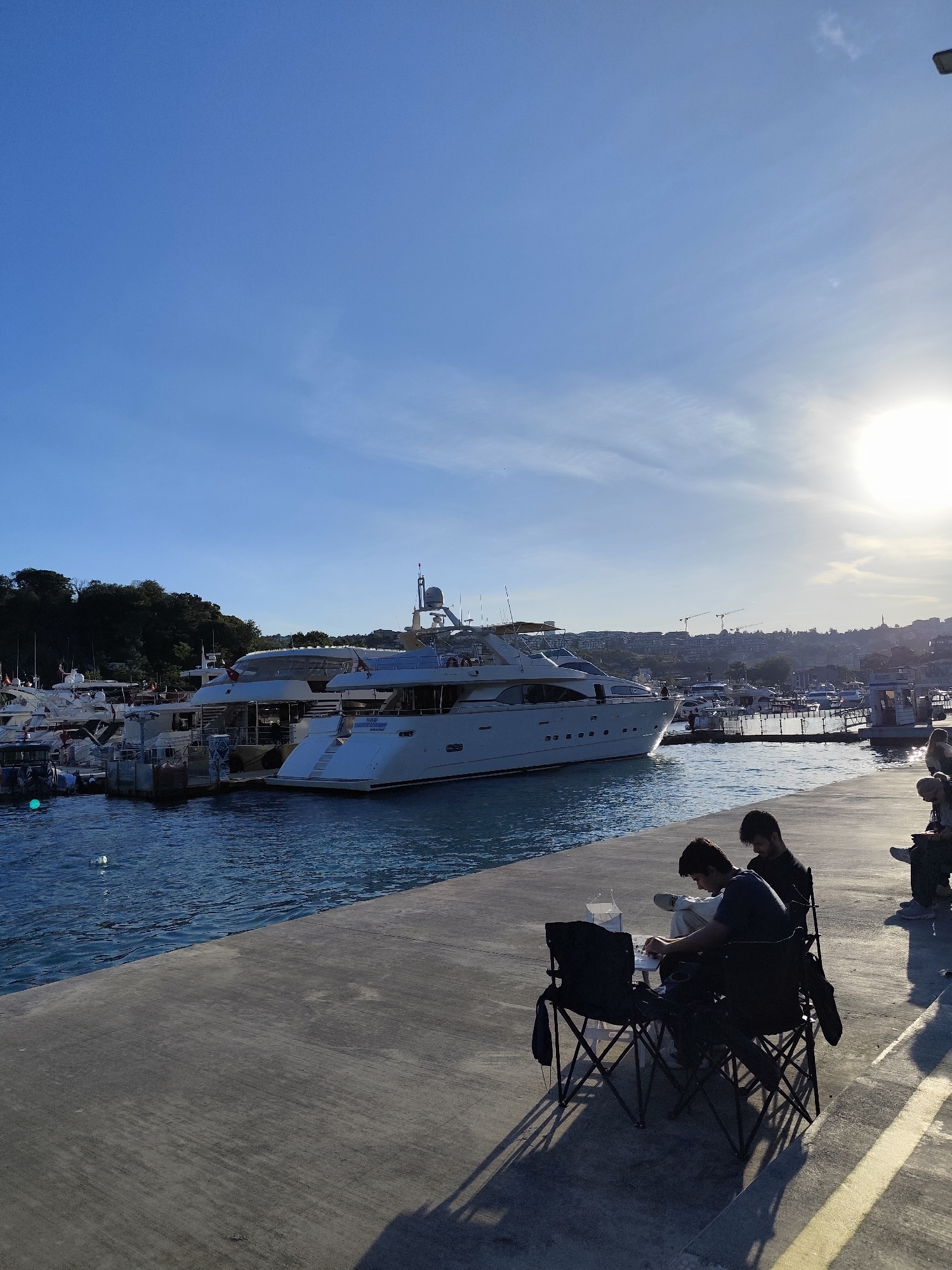 The image depicts a marina in Sarıyer, Turkey, under a bright, clear sky with the sun positioned to the right, causing glare. Numerous yachts and smaller boats are docked in the marina. A large, multi-deck yacht is prominently featured, docked along a concrete pier.
On the pier, two men are seated in folding chairs, engaged in a chess game. Other individuals are seated further down the pier, closer to the right edge of the frame. The environment is a mix of natural and urban elements, with trees visible in the background on the left and buildings on the horizon. The pier is made of gray concrete.