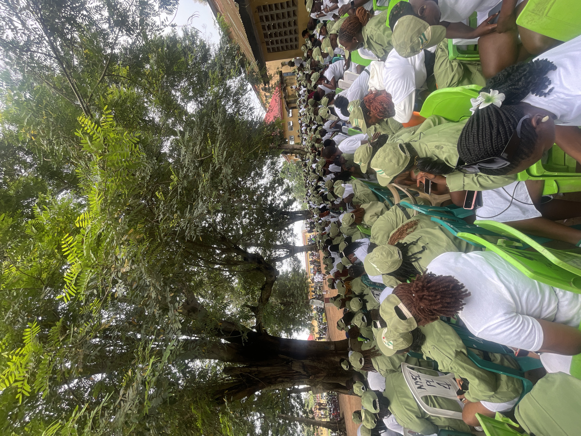 A medium-shot, high-angle, vertical photograph captures a large gathering of people, predominantly young adults, seated outdoors in Emure-Ekiti, Nigeria. The scene is dominated by a massive, ancient tree with a thick trunk and extensive canopy of green leaves, providing shade over a significant portion of the assembled crowd. Many individuals are wearing matching light green uniforms and caps, suggesting an organized event or program, possibly a youth service orientation given the "NYSC PL 4" visible on a white plastic chair in the foreground.
The attendees are seated on green and white plastic chairs arranged in rows. The individuals in the foreground are facing away from the camera, while those further back are partially visible. The environment appears to be a clearing or courtyard with buildings constructed from yellow brick visible in the background, some with corrugated metal roofing. The ground is dirt or sand. The atmosphere seems to be one of attentive waiting, with some individuals looking towards an unseen focal point in front of them, possibly a stage or speaker. The lighting suggests it is daytime, with diffused sunlight filtering through the tree canopy.