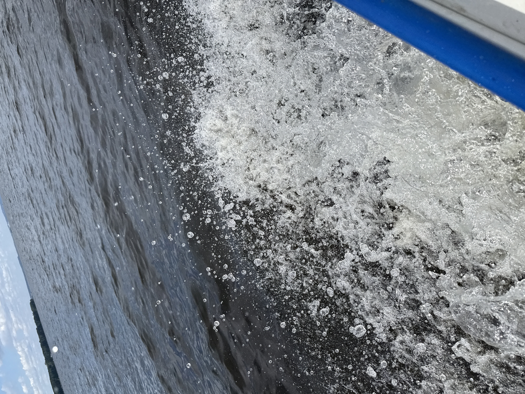 This image, captured in Chaikovsky, Russia, presents a dynamic, close-up perspective of water being churned by a moving boat. The majority of the frame is filled with the turbulent interaction of water, characterized by an abundance of white spray, foam, and countless shimmering water droplets. These droplets, varying in size, are suspended against the darker, deeper water, creating a stark contrast and highlighting the energetic motion.
In the upper right corner, a segment of a blue railing or boat edge against a white surface is visible, firmly placing the viewer's perspective on board a vessel. The water nearest this edge is intensely agitated, frothing into dense white masses and larger splashes. Moving away from the immediate impact zone, towards the left of the frame, the water transitions to a darker, rippled surface, indicating the wake created by the boat's passage.
The background, particularly on the far left, reveals a distant, low-lying shoreline or island covered with dark green foliage under a bright sky with scattered clouds. This suggests an open body of water, likely a river or large reservoir, consistent with the location context of Chaikovsky (e.g., Kama River or Votkinsk Reservoir). The clear skies and bright illumination confirm it is daytime. The overall impression is one of rapid movement across the water. There is no visible text in the image.