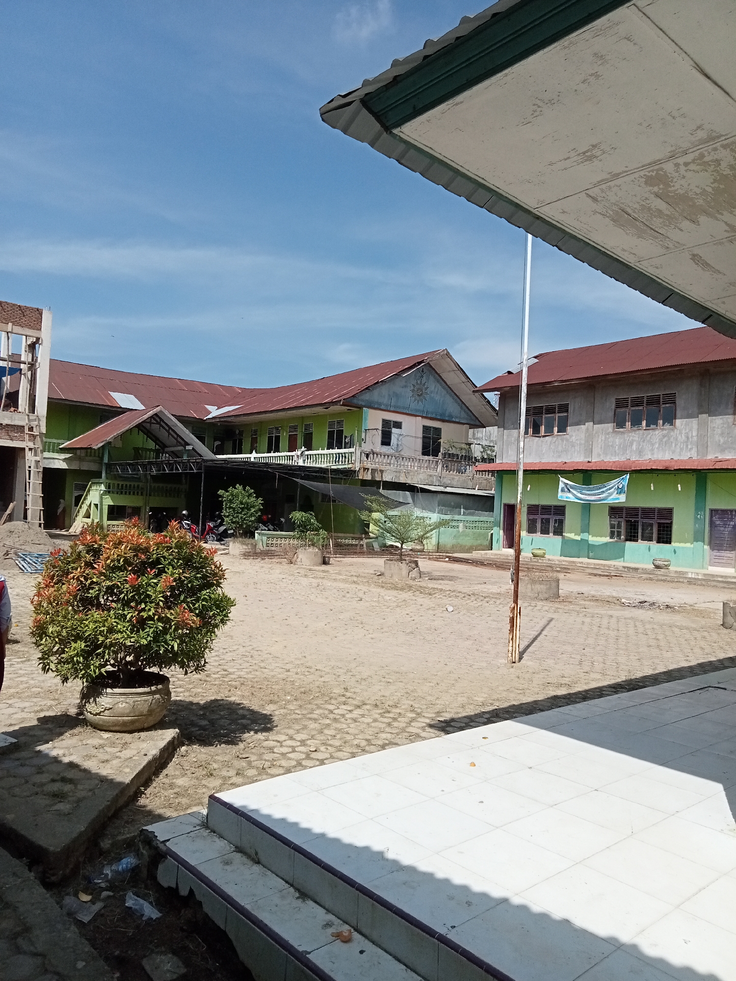 The image depicts a view of a school courtyard in Kutalhoksukon, Indonesia, under a clear blue sky. In the foreground, a large potted plant with red-tipped leaves is visible on a concrete slab. To the right, a white tiled staircase leads up, with shadows cast across it. The courtyard itself is paved with interlocking concrete blocks and has a sandy texture.
In the mid-ground, a multi-story building with green walls and a red roof dominates the scene. It has a veranda with railings on the upper floors and covered walkways on the ground level. Several motorcycles are parked under the covered area. Adjacent to this building is another structure with gray walls and a red roof, featuring rows of windows. A banner, partially visible, hangs from this building. A flagpole stands in the courtyard between these buildings.
Further in the background, to the left, a partially constructed building with scaffolding is visible. The environment appears to be a typical school setting, with educational buildings and outdoor space for students. The overall impression is of a sunny day at a school in a tropical or subtropical region.