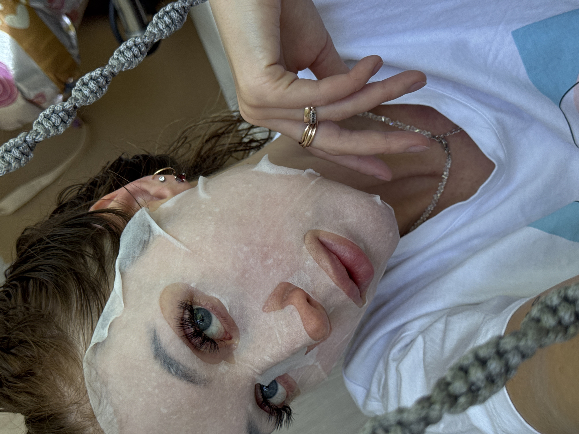 The image shows a woman lying down, possibly in an indoor setting in an unknown city. She has a sheet mask on her face. The woman is wearing a white t-shirt and a necklace. She is wearing rings on her fingers. A decorative rope-like object is visible in the foreground. The exact location in the city of Chaikovsky, Russia is undeterminable.