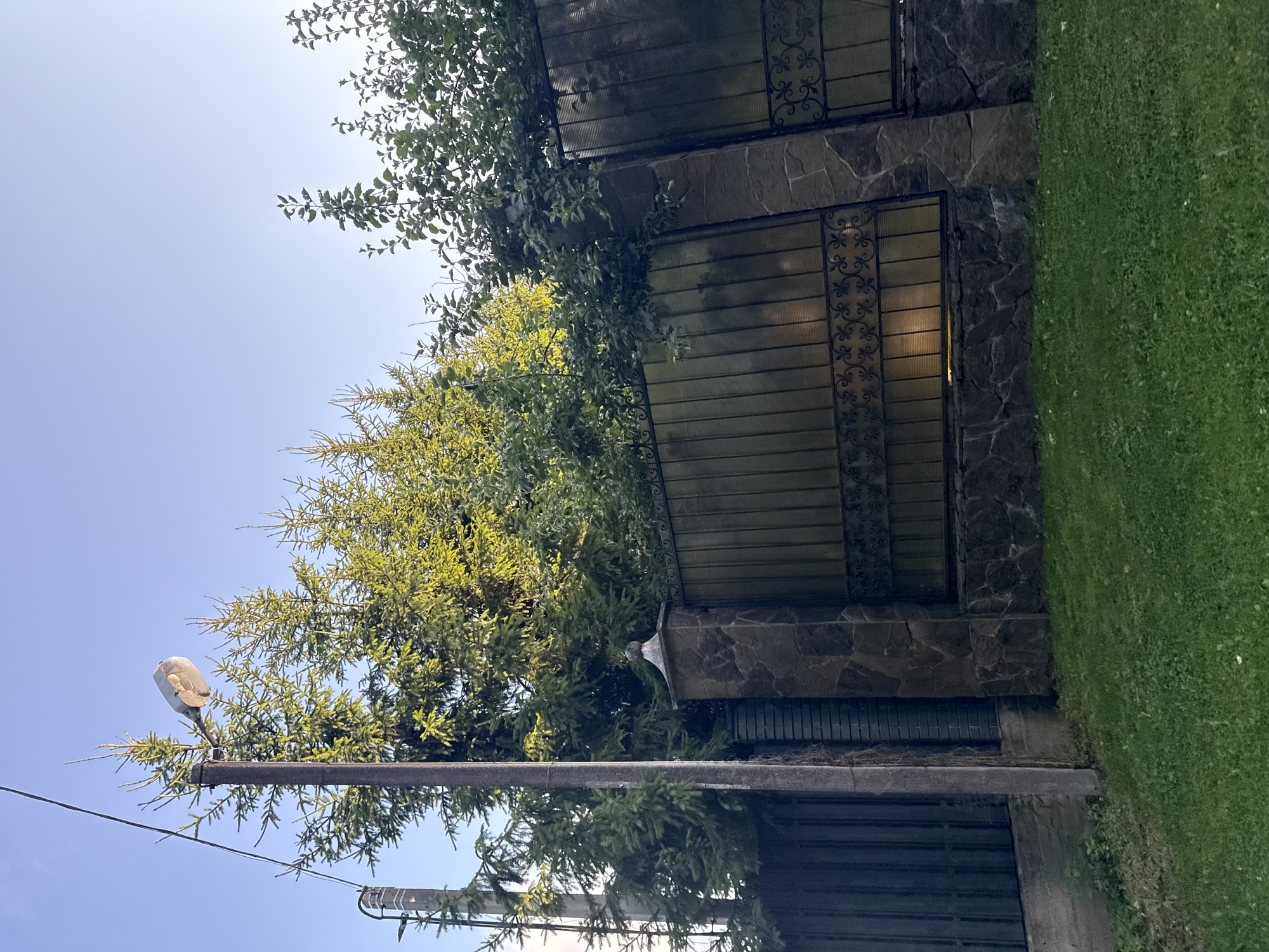 The image depicts a residential fence in Yukki, Russia, on a clear day. The fence is constructed with dark metal panels and decorative wrought iron accents, supported by stone pillars. Behind the fence, a line of evergreen trees and deciduous trees are visible, some of which are illuminated by sunlight. A street light is mounted on a utility pole to the left of the fence, with power lines extending overhead. The foreground shows a well-maintained green lawn. There are no people or animals present in the scene.