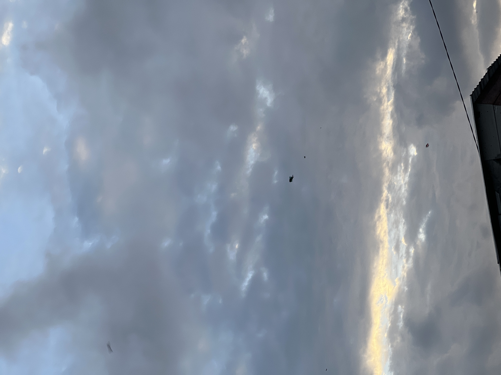 The image depicts a cloudy sky with a hint of sunlight breaking through the clouds. In the distance, several kites can be seen flying. The lower portion of the image shows the roof of a building. The scene is set in the city of Mengwi, Indonesia, where cloudy skies are common. There are no people or animals visible in the image.