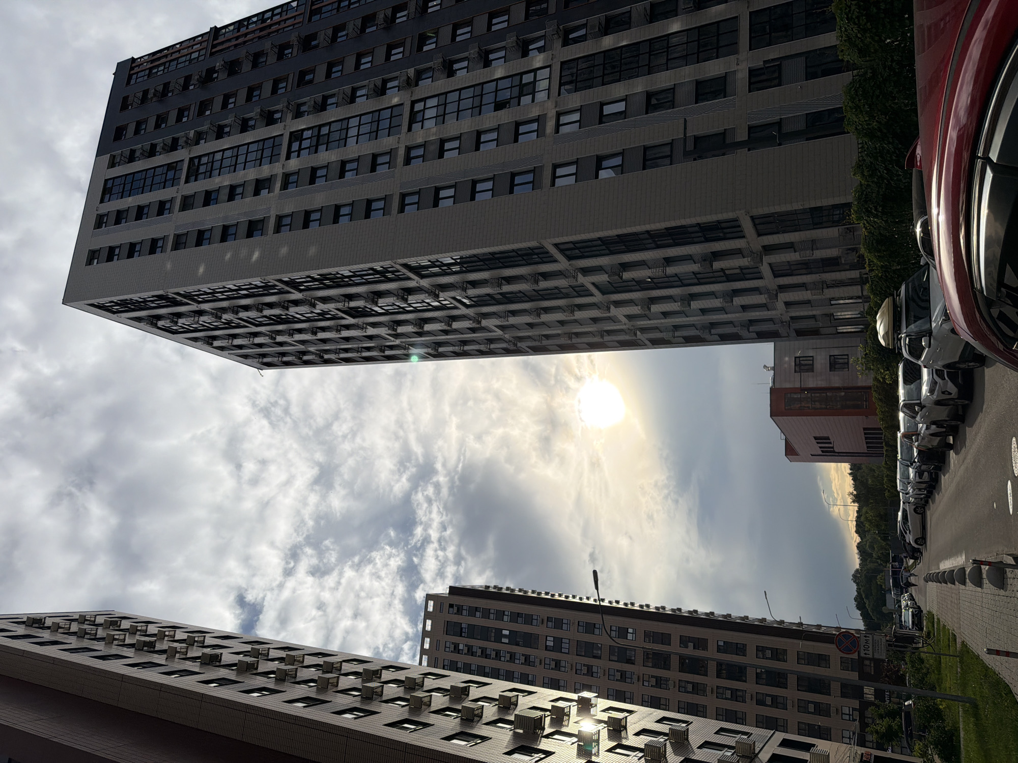The image depicts a street scene in a modern residential area, likely on the outskirts of Moscow, Russia, characterized by high-rise apartment buildings. The sky is partially overcast with the sun visible, creating a bright spot and lens flare in the upper right quadrant. Two prominent, tall apartment buildings dominate the frame, one on the left and a larger one on the right. Both buildings feature numerous windows and external air conditioning units.
In the foreground and mid-ground, a street is lined with parked cars. A sidewalk with decorative concrete bollards runs along the left side of the street. A street lamp is visible. Further down the street, more cars are parked, and a few pedestrians are walking. A smaller, brick-colored building is visible behind the parked cars on the right. Trees and green foliage line the background, suggesting a suburban or newly developed urban environment. A red car is partially visible in the extreme foreground. A traffic sign with a red circle and a horizontal line is mounted on a pole. The overall impression is of a contemporary urban landscape under a cloudy sky.