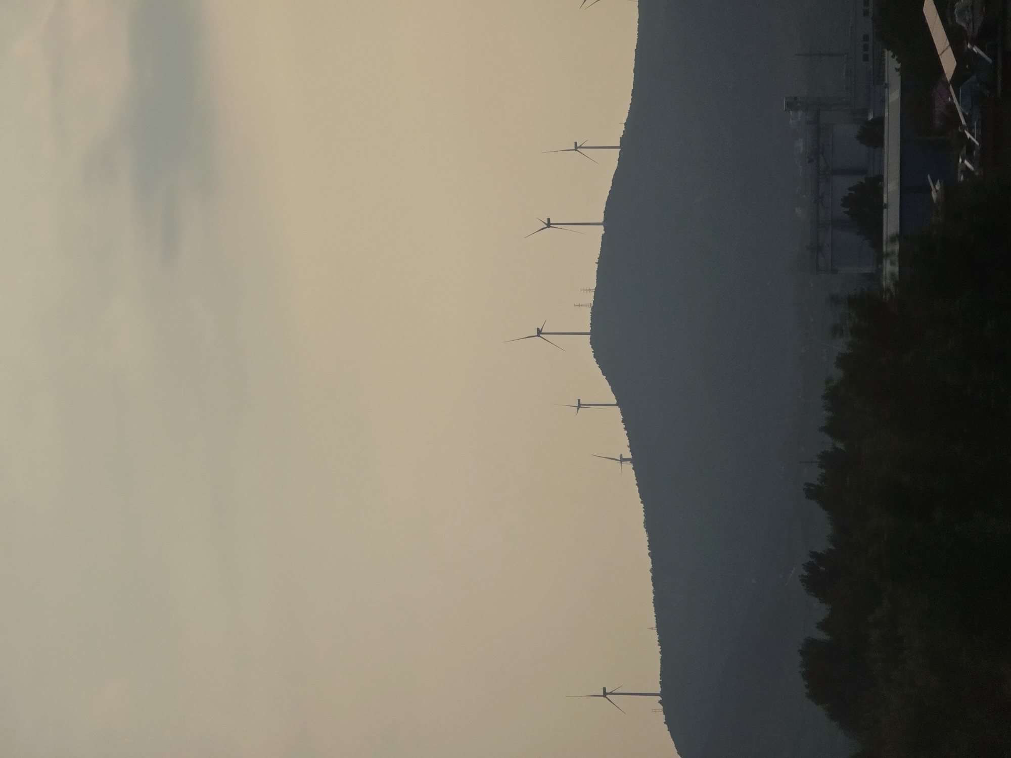 A silhouetted mountain range is covered in wind turbines. The sun is setting or rising and casting a hazy glow on the sky. The turbines are spaced out along the ridge of the mountain, with their blades extended. Some appear to be in motion, while others are still. The scene is peaceful and serene, evoking a sense of natural power and sustainability. In the foreground, there are trees, and in the distance, a factory or industrial complex can be seen. The lighting suggests it's either dusk or dawn.