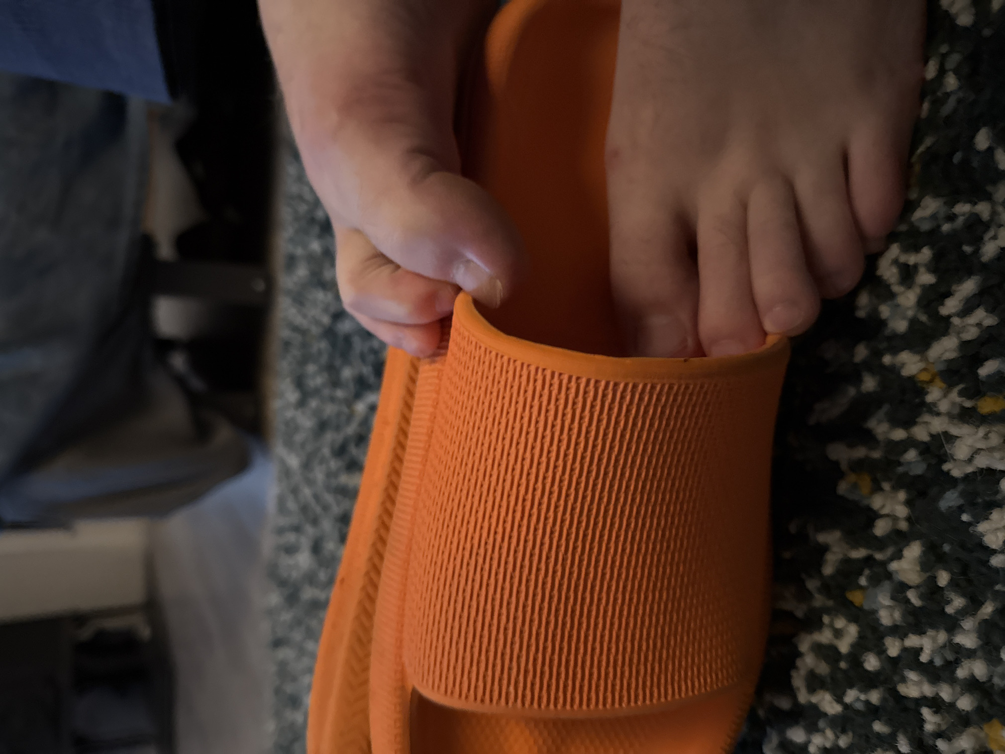 In a close-up, top-down view, a person's right foot is being inserted into an orange slide sandal. The sandal, made of a textured synthetic material, is positioned on a dark, flecked carpet. The foot is pale and appears to be that of an adult, with visible toes and toenails. The individual's hand is visible, guiding the foot into the sandal. The background is blurred, showing a dark, upholstered piece of furniture. The overall lighting is dim.