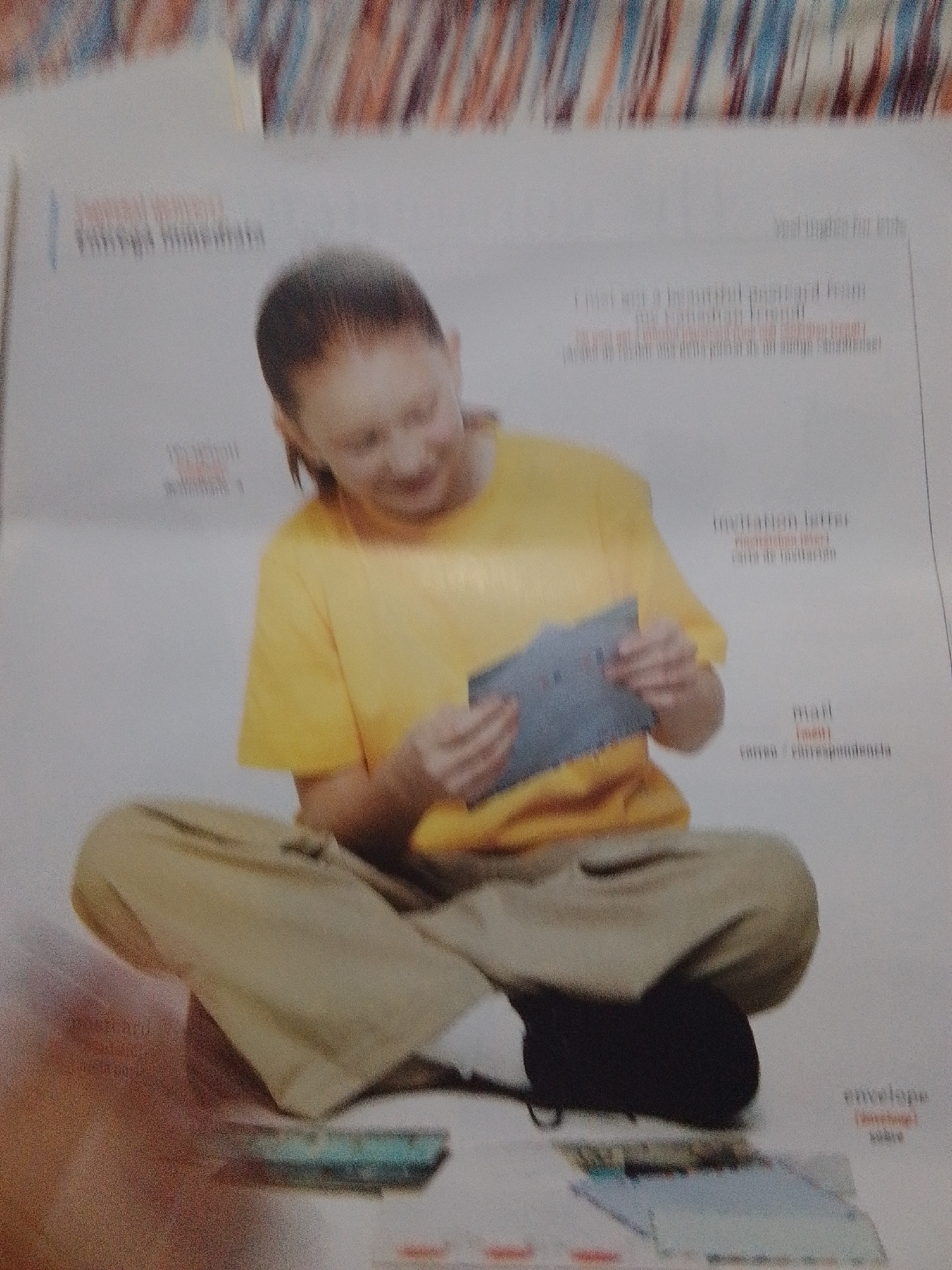 The image shows a young woman sitting on the floor, holding an envelope and a card, possibly engaged in writing a letter or sending mail. The text in the image refers to postcards, letters, and envelopes, indicating a theme related to communication and correspondence. The scene appears to be indoors, with a blurred background that suggests a domestic or educational setting. There is no visual information in the image that provides details about the city of La Tagua, Venezuela.