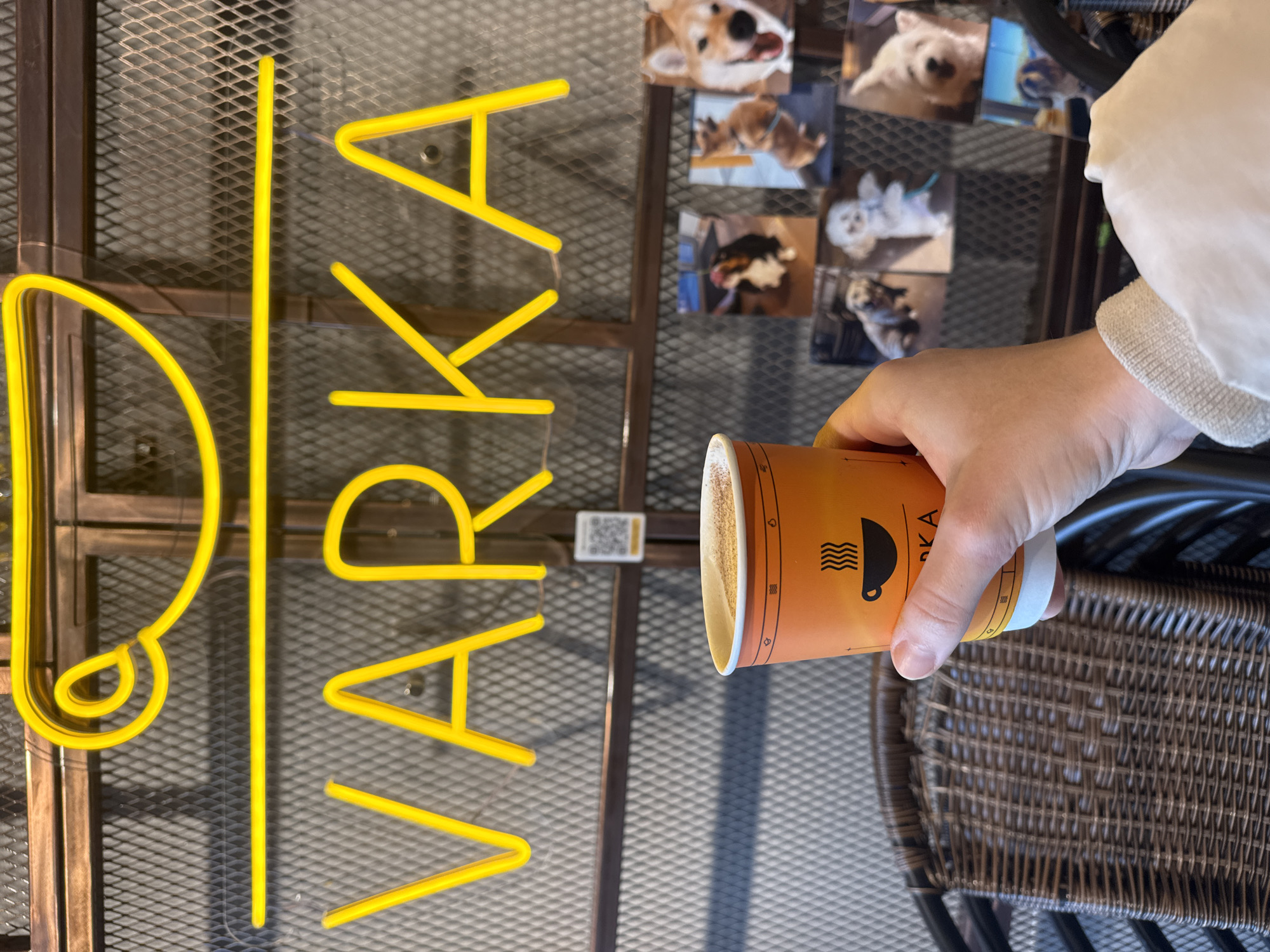 The image shows a person holding an orange coffee cup in front of a neon sign that spells "VARKA" and a stylized coffee cup. Behind the sign, there is a wall with a diamond-patterned mesh and several framed pictures of dogs. The scene appears to be indoors, possibly a cafe or a similar establishment, with a wicker chair visible in the foreground. The overall lighting suggests a daytime setting. The coffee cup has a logo that includes a steaming coffee icon and the letters "VARKA" below it. The neon sign is bright yellow. The pictures of dogs include various breeds and poses.