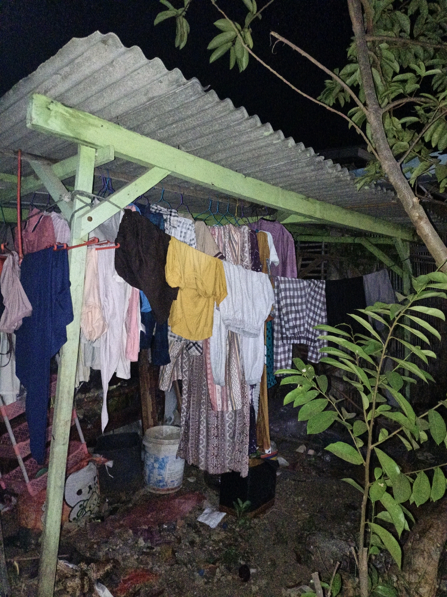 An outdoor scene at night shows a clothesline with various items of clothing hanging on it, underneath a corrugated metal roof supported by a green wooden structure. The ground is covered with debris, leaves, and dirt. To the right of the clothesline, a tree with green leaves is visible. The image appears to be taken in a somewhat unkempt outdoor area, possibly a backyard or alleyway, in Cikal, Indonesia. There are no people visible in the image. The scene suggests a functional setup for drying laundry, with the surrounding environment appearing somewhat neglected.