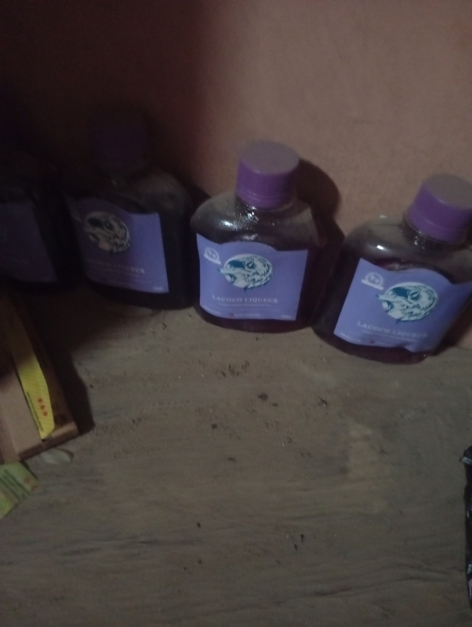 The image displays four bottles of "LaCoco Liqueur" standing upright on a gritty surface, possibly dirt or sand. The bottles have purple caps and labels featuring a circular logo with a depiction of what appears to be a wild animal, possibly a dog or wolf, and text. The background is a plain, light-colored wall. The lighting is dim, and the image quality is somewhat blurry, suggesting it was taken indoors with poor lighting conditions. There are no people visible in the image, and no discernible activities or interactions are taking place. The scene is static and appears to be a simple arrangement of the bottles. The location within Jalingo, Nigeria, cannot be determined from the visual information provided in the image.