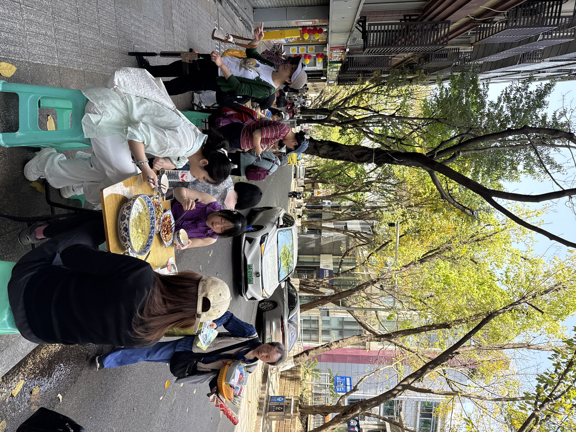 The image shows a street scene in the Yuzhong District of China, with people seated at tables and eating outdoors. They are seated on low, green plastic stools. The tables are small and have bowls of food. The food appears to be noodles and peanuts. The people are dressed in casual clothing. A white electric vehicle can be seen parked nearby with license plate WA D18582. Trees line the street, casting shadows. Buildings are visible in the background. There are garbage cans along the roadside.