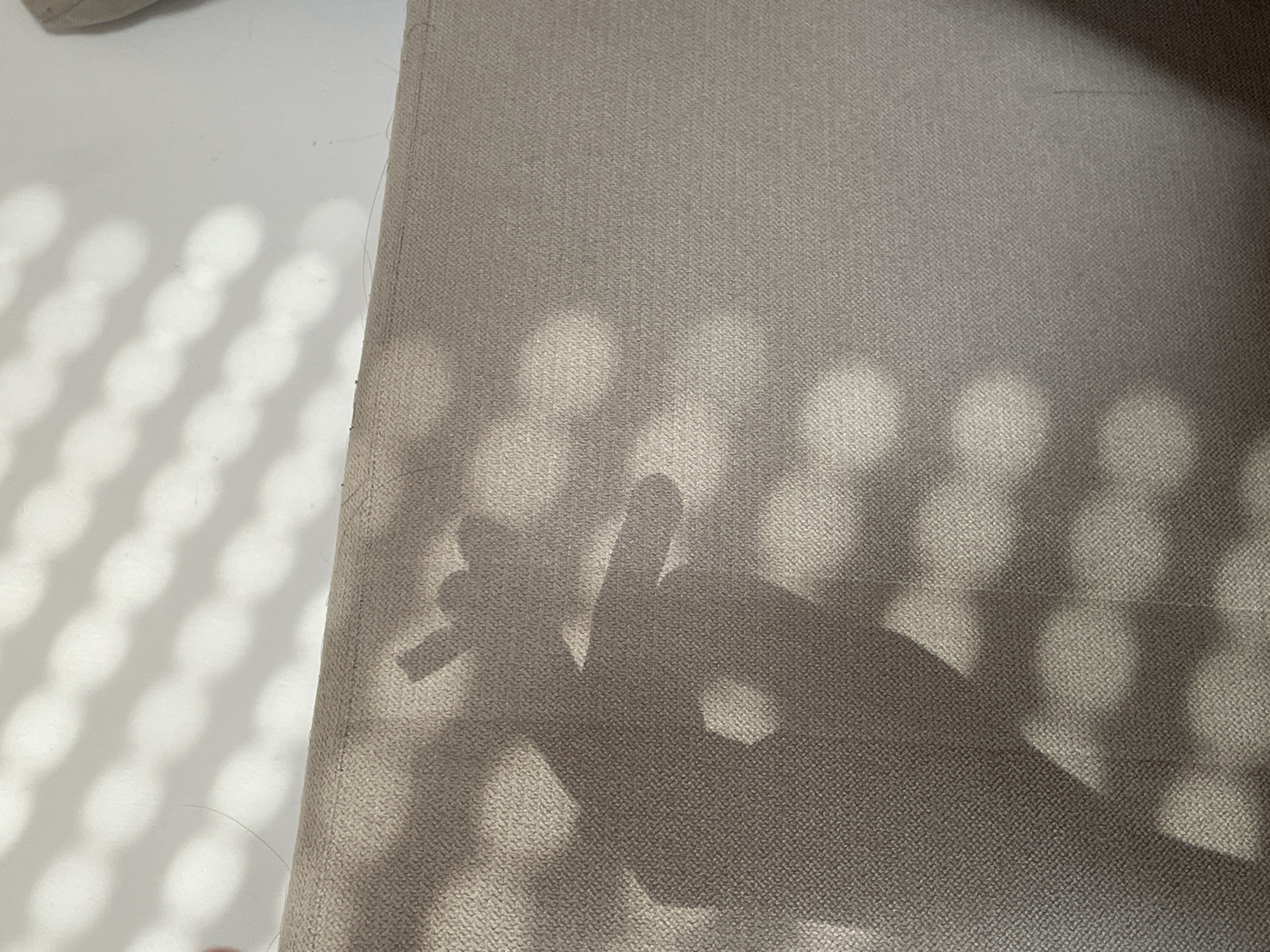 The image shows a textured, light brown or beige surface, possibly fabric or upholstery. There are strong shadows cast across the surface, creating a pattern of rounded shapes. A shadow of a hand making a peace sign is partially visible on the surface, which might suggest the setting is an interior where light from a window or another source is casting the shadows. The location in Belgrade, Serbia, cannot be determined with the information provided.