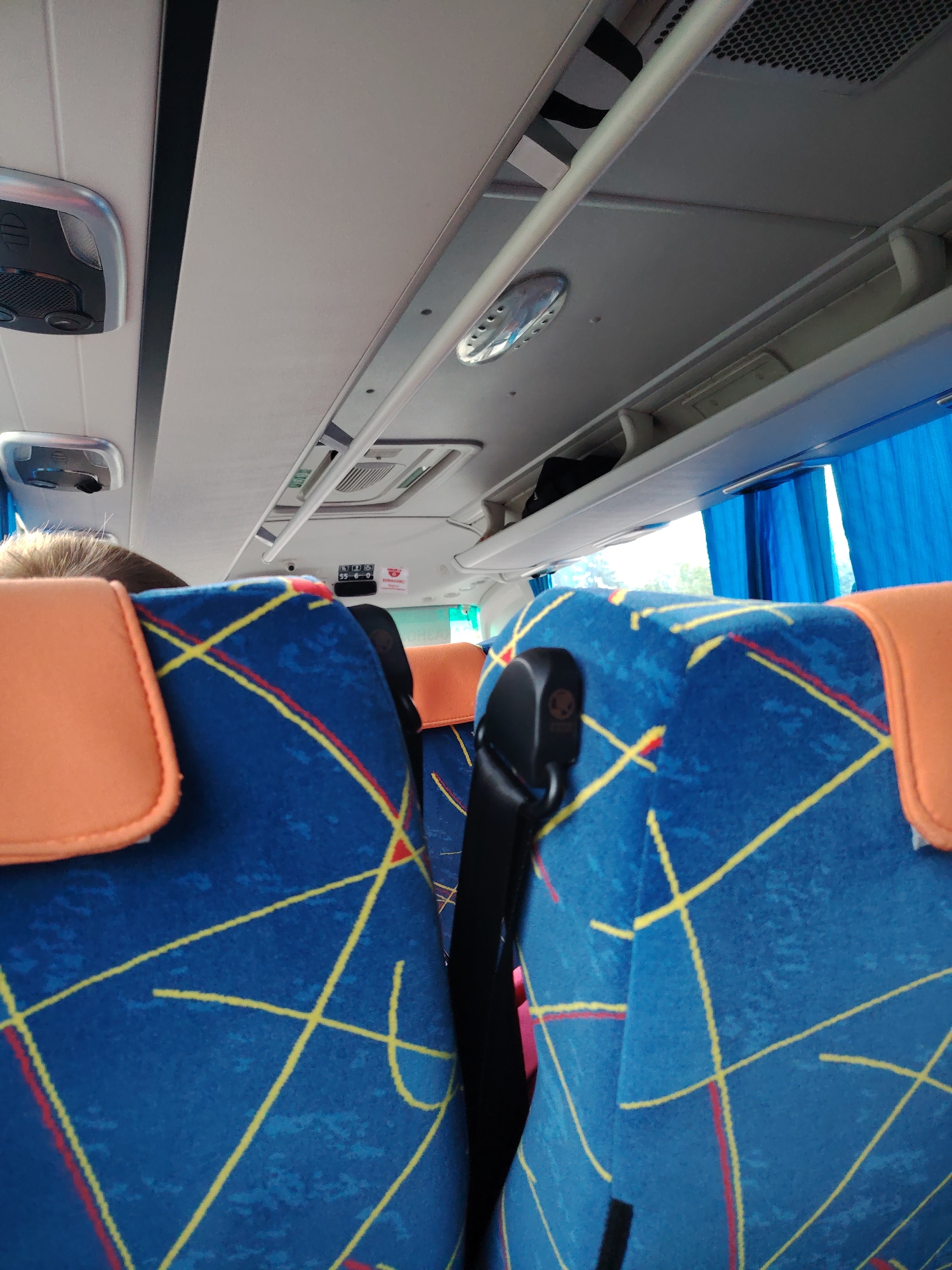 The image shows the interior of a bus, looking forward from the perspective of a seated passenger. Two blue bus seats with orange headrests are visible in the foreground, both featuring a geometric pattern of red and yellow lines. A black seatbelt is attached to the right seat. Above the seats, the bus's ceiling is visible, with overhead compartments, lighting fixtures, and air vents. Blue curtains are drawn across the windows on the right side of the bus. The scene is likely set within the city of Konakovo, Russia, as indicated by the context. There are no people clearly depicted, only the back of a passenger's head is visible on the left. The environment is clean and appears to be in good condition.