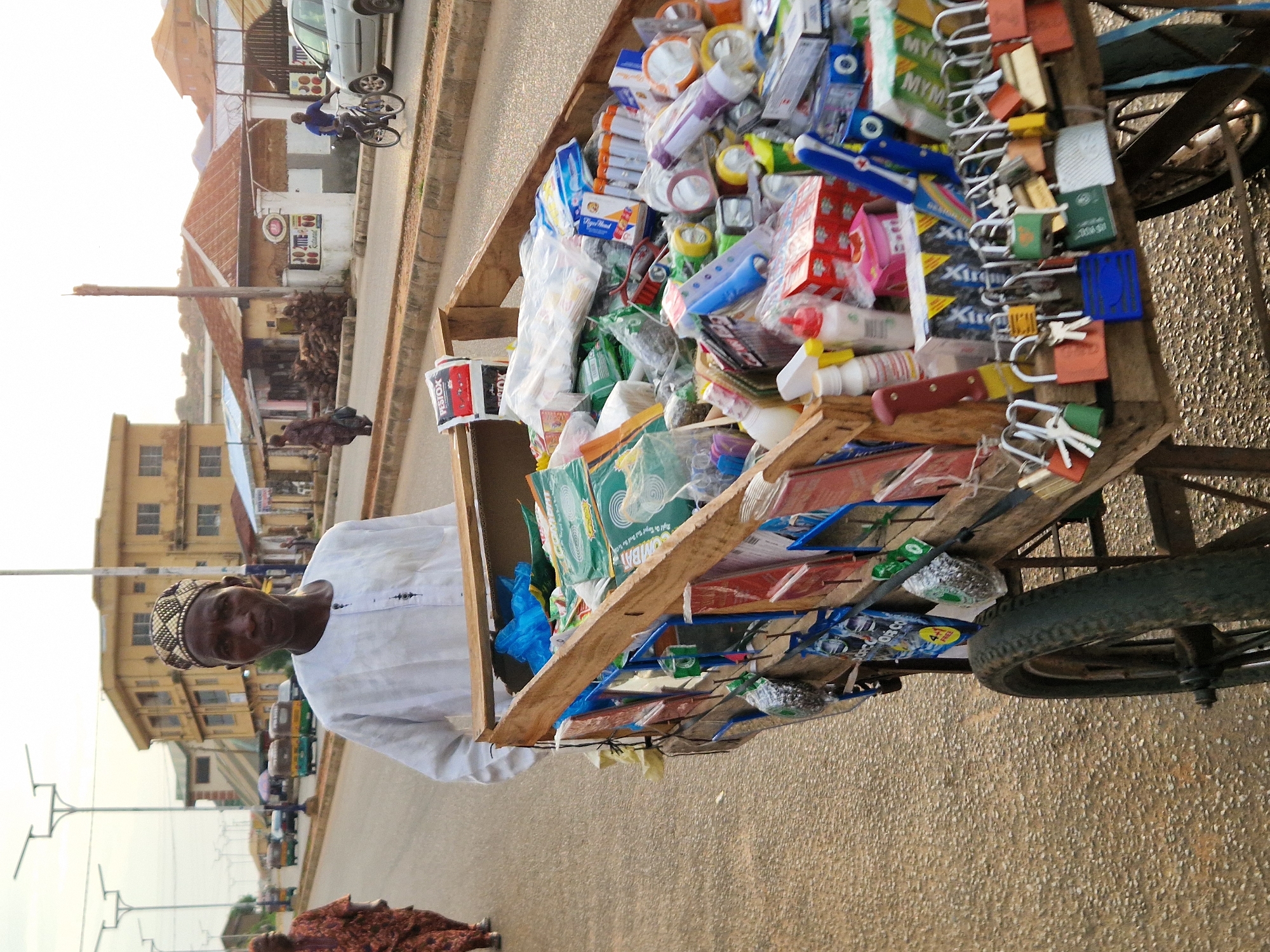 A man in a white traditional outfit and patterned cap pushes a wooden cart filled with various items, including locks, batteries, and packaged goods, down a paved street in Kabong, Nigeria. The street is lined with buildings and utility poles, and other pedestrians and vehicles are visible in the background. The man is the central figure, walking towards the viewer and pushing the cart with both hands. The cart is laden with a diverse assortment of merchandise, suggesting he is a street vendor. The environment appears to be an urban setting with a mix of commercial and residential structures.