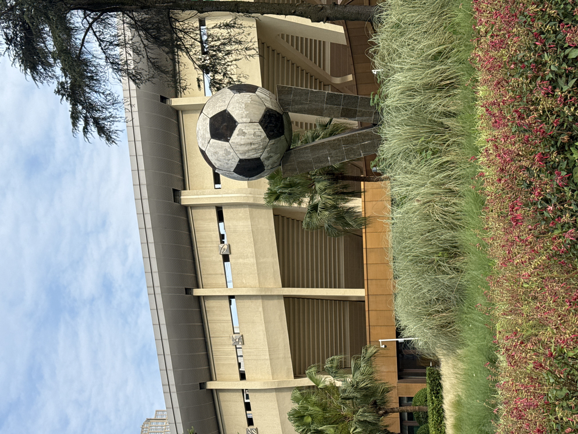 The image shows a large soccer ball sculpture in front of a tall, modern building. The building has a multi-tiered design with multiple columns. The sculpture is supported by a geometric base. Lush greenery surrounds the sculpture, and a tree branch is visible in the upper left corner. The sky is blue with some clouds. It is in the city of Qingyang District, China.
