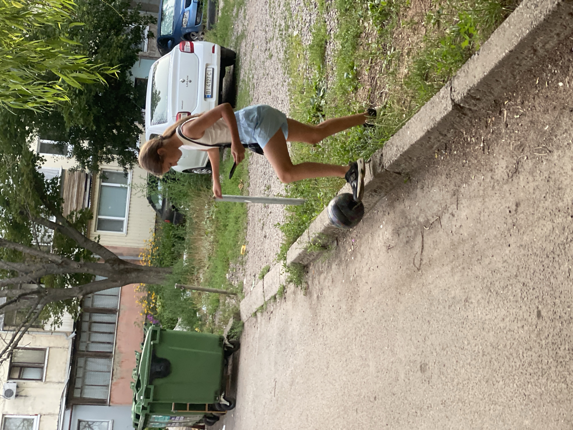 In Poltava, Ukraine, a young girl with light brown hair pulled back in a ponytail, wearing a white tank top and light blue shorts, is standing on a concrete curb. She is holding a black smartphone in her right hand and a black basketball is at her feet, positioned on the curb. Her left leg is bent at the knee, and her right leg is extended. The girl is facing towards the left of the frame. Behind her, a white sedan with a license plate that reads "BI 4127 CI" is parked. To the left of the girl, a tall, thin metal pole is visible. In the background, there is a multi-story building with balconies and trees. Further to the left, two large green refuse bins are situated. The ground is a mix of asphalt and gravel, with patches of green grass and weeds growing around the curb and in the background. The lighting suggests it is daytime, possibly overcast.