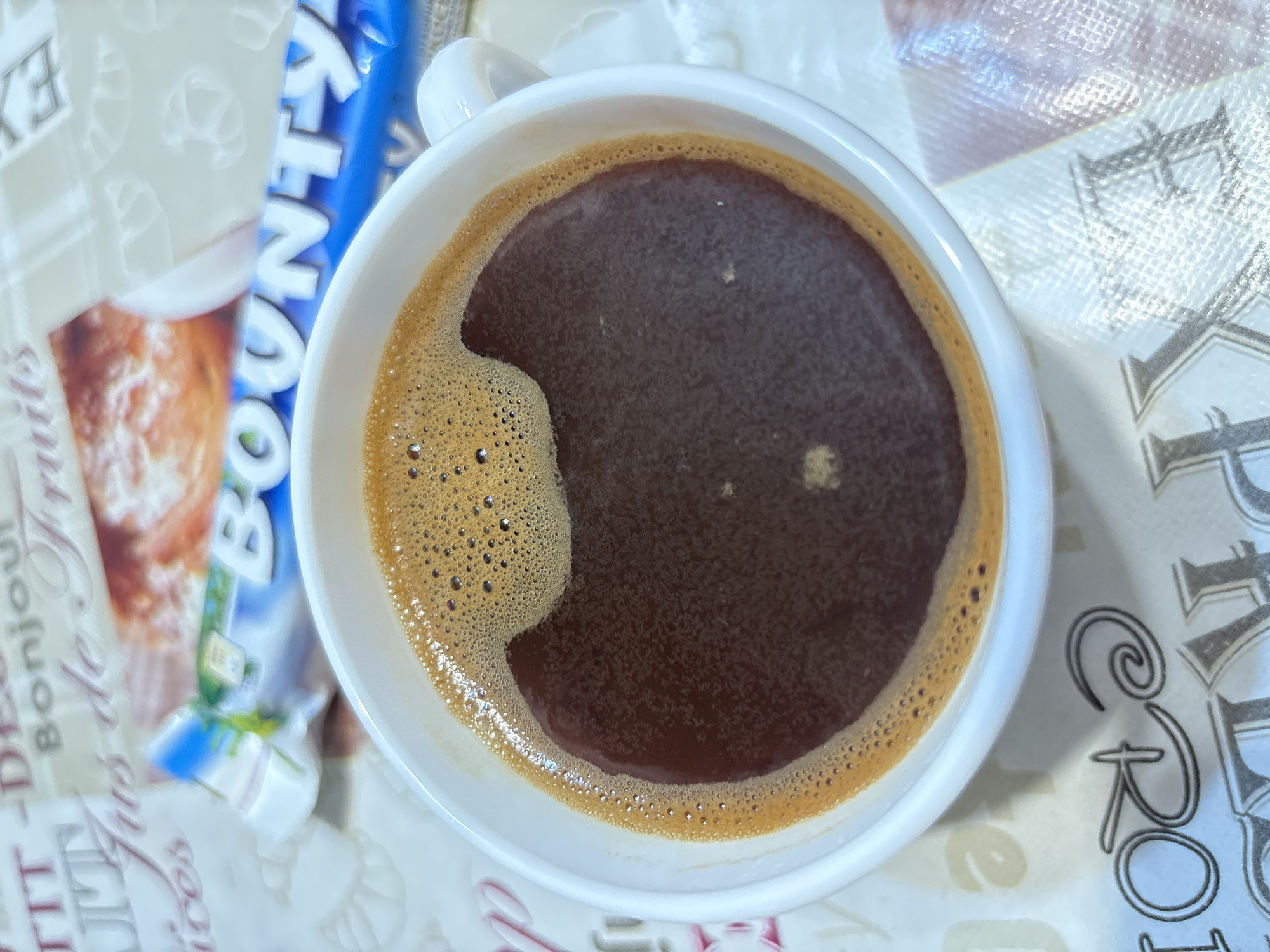 The image shows a close-up overhead view of a white mug filled with dark coffee, topped with a layer of foam. The coffee has a rich brown color and the foam is a lighter tan with small bubbles. A wrapper for a "Bounty" candy bar is visible in the background, partially obscuring the mug and resting on a patterned surface. The surface appears to be a tablecloth or placemat with various words and decorative elements, including some French text and ornate lettering. The lighting suggests it is indoors, possibly in a home or cafe setting. There are no people or animals in the image. The location in the city of Zubov, Russia is not determinable from the visual information provided in the image.