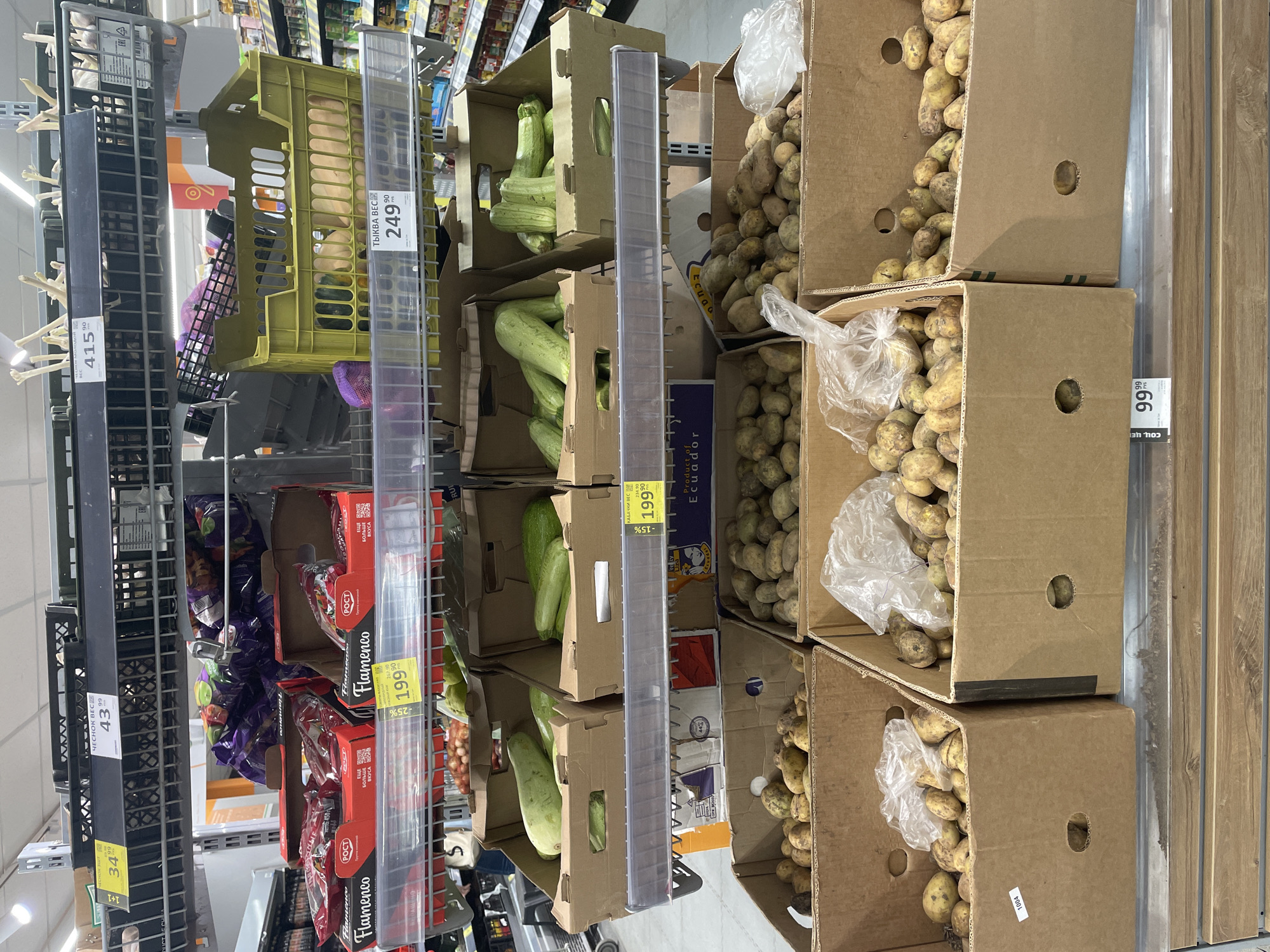 This is a medium shot, rotated 90 degrees clockwise, of a grocery store aisle filled with produce. The shelves are laden with various vegetables and some bagged items. In the foreground, on the left, are black plastic crates containing what appear to be garlic bulbs and purple net bags of onions. A price tag indicates "41590". Next to these, a bright yellow plastic crate holds what looks like squash or gourds, with a price tag of "24990" and text reading "ТЫКВА ВЕС" (Pumpkin Weight).
Moving to the right, stacked cardboard boxes display zucchini. One box has a price tag of "19990" and "-15%". Another stack of boxes contains potatoes, with some in clear plastic bags. A price tag for the potatoes reads "9999". Further to the right, more cardboard boxes hold potatoes. There are also red boxes with "Flamenco" branding, likely containing some type of processed food or seasoning, with price tags like "19990" and "-25%". Another price tag indicates "4399" for "ЧЕСНОК ВЕС" (Garlic Weight).
The setting is clearly a supermarket aisle, with bright overhead lighting. The overall impression is one of abundance and variety in the produce section of a store. The time of day cannot be determined.