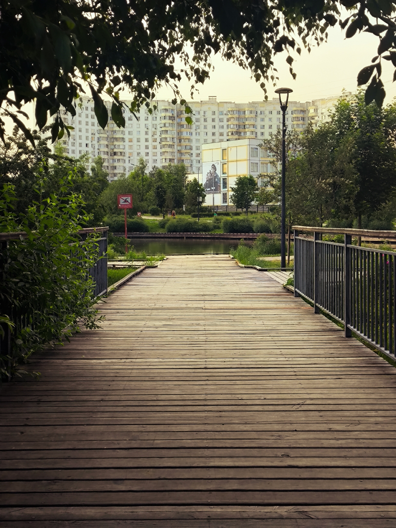 A wooden boardwalk curves towards a calm body of water in a park setting. Green foliage frames the left side of the boardwalk, while a black metal railing lines the right. Beyond the water, a street lamp stands beside a large apartment building with numerous windows. A sign with a red border and a graphic of a person jumping into water is visible on a pole to the left of the water. The sky is overcast and pale. The scene is outdoors during the daytime.