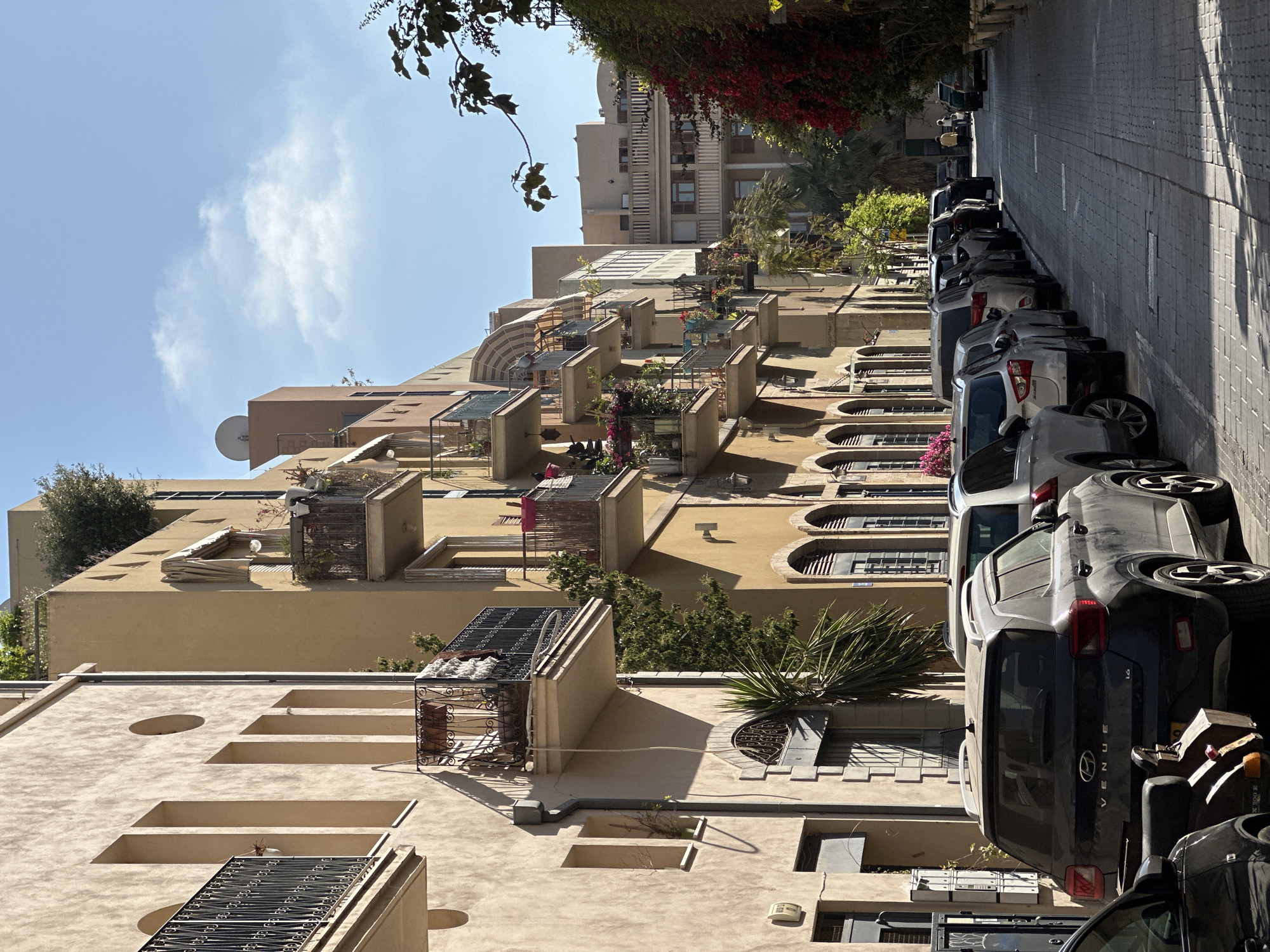 The image displays a bright, sunny street scene, rotated 90 degrees clockwise, depicting a residential area consistent with Tel-Aviv, Israel.
The main subjects are a long row of multi-story residential buildings on the left and a parallel street lined with parked cars on the right. The buildings are primarily constructed from light beige or sandy-colored materials, suggesting plaster or stucco. They exhibit a mix of architectural styles, featuring numerous balconies adorned with various plants and small trees. Some balconies have decorative railings and awnings, while others appear more open. Arched windows are visible on the lower floors of some structures, contrasting with more rectangular openings on higher levels. Rooftops are visible, featuring satellite dishes, various structures, and additional greenery. Red flowering vines cascade over some of the upper building sections, adding a splash of color.
The street itself is paved with light-colored, patterned blocks or cobblestones, and it appears relatively narrow, likely a one-way street. A dense line of cars is parked bumper-to-bumper along the curb. The vehicles are a mix of sedans and SUVs, predominantly in shades of grey, silver, and black. A prominent vehicle is a dark grey Hyundai Venue, clearly showing its model name "VENUE" and "1.6" on its rear trunk. Other car models are less discernible but indicate common modern vehicles.
The activity taking place is primarily the static scene of parked cars along a residential street. No people are visible, and there are no active events depicted.
Notable details include the bright, clear blue sky with a few wispy white clouds, indicating excellent weather. Strong, well-defined shadows cast by the buildings and cars suggest the sun is high overhead, likely midday or early afternoon. The overall atmosphere is calm and quiet, characteristic of a residential urban environment during the day.