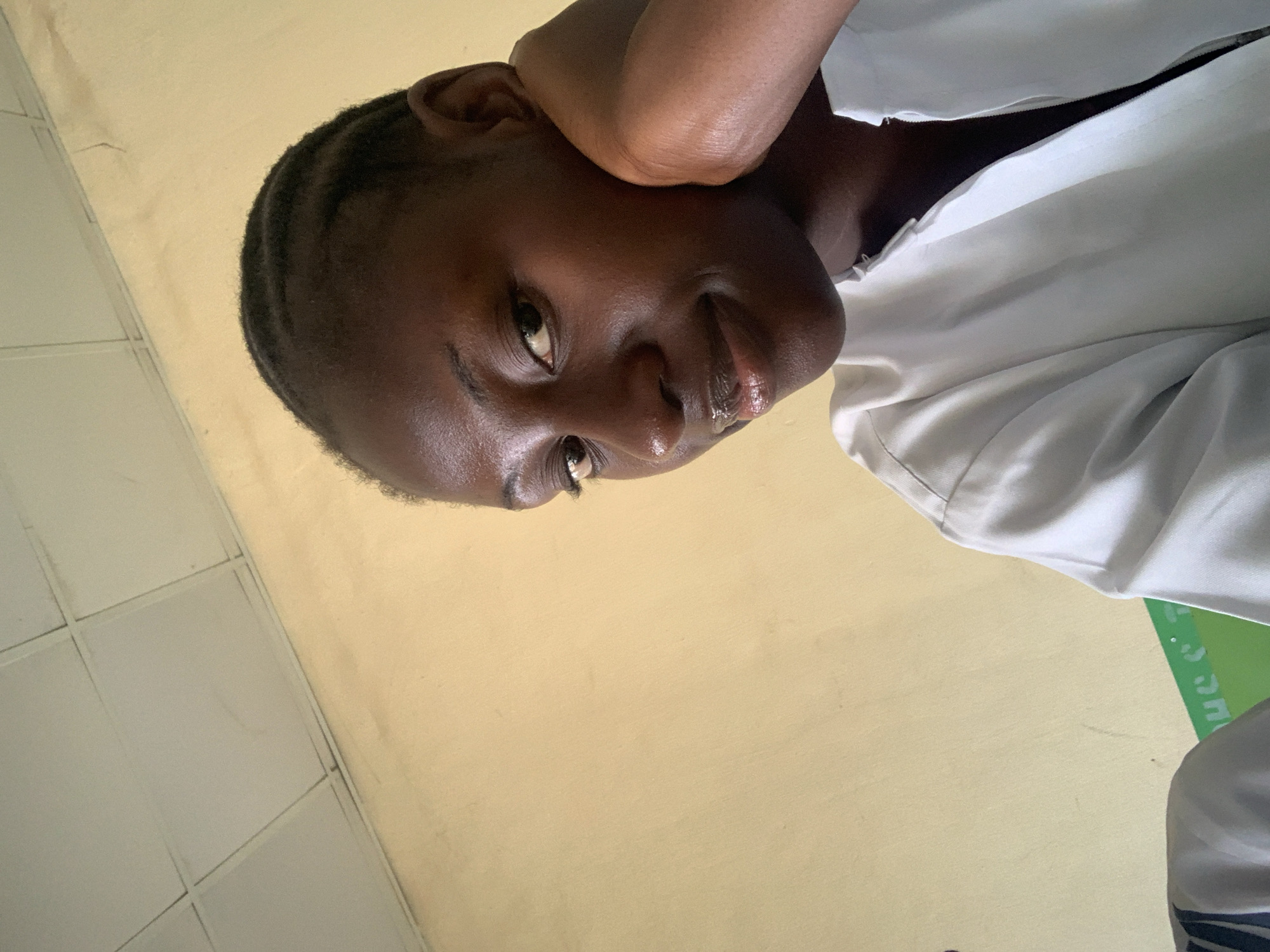 A close-up, slightly tilted selfie of a young Black woman with short, braided hair. She is wearing a white collared shirt. Her right hand and elbow are raised and positioned near her head. The background includes a wall with light-colored tiles and a section of a light yellowish painted wall. It is unknown if this image was taken in Keffi, Nigeria.