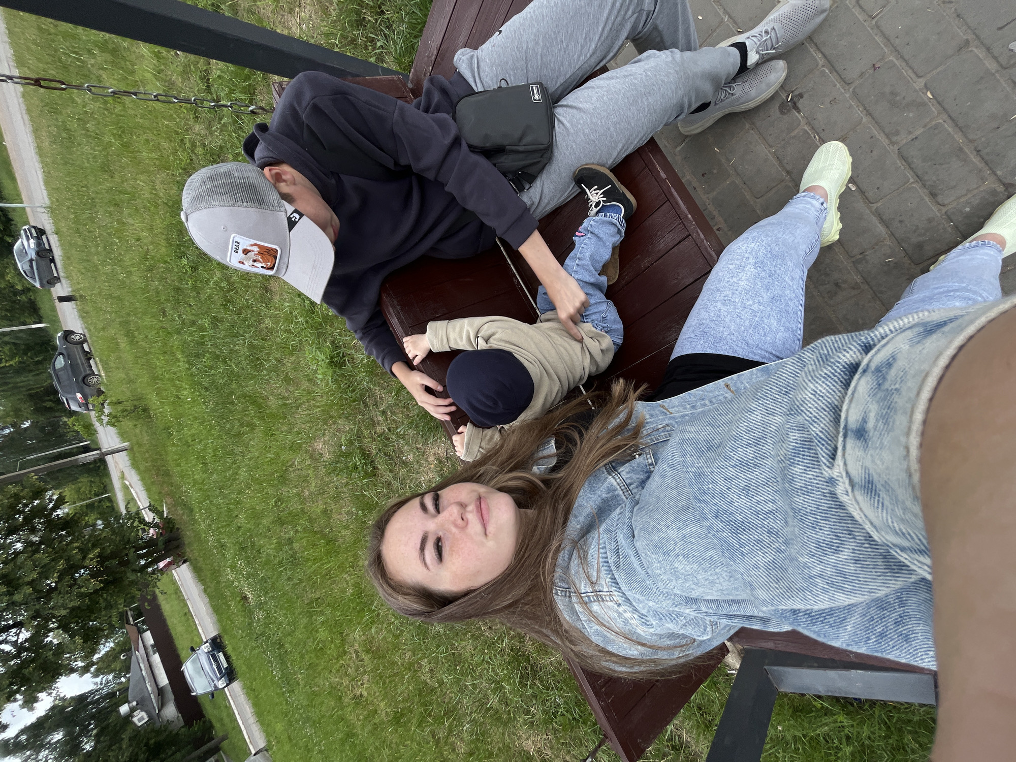 In an outdoor setting, possibly a park, in the city of Chaikovsky, Russia, a young woman with long brown hair and freckles is taking a selfie. She is wearing a light blue denim jacket. Next to her on a wooden swing bench sits a person wearing a grey baseball cap with a patch that says "BEAR," a dark hoodie, and grey sweatpants. This person is holding a young child who is wearing a light beige jacket and blue jeans. The child is sitting on the bench between the two adults. The background shows a grassy area and a road with cars.