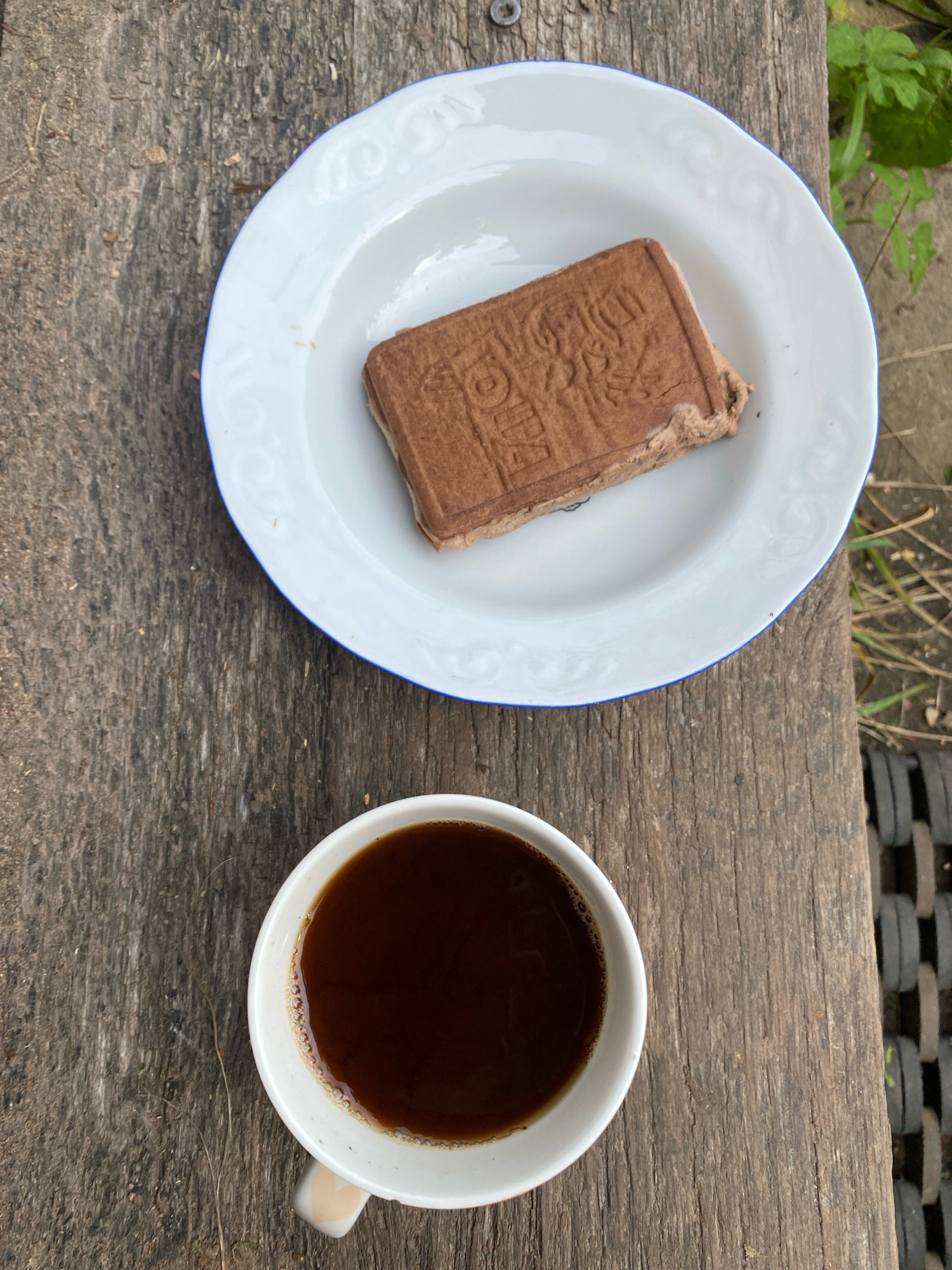 A white plate with a blue rim sits on a rustic wooden surface. On the plate rests a rectangular chocolate confection with embossed lettering and a textured surface. Below the plate, a white mug filled with dark coffee is also on the wooden surface. The background consists of dried grass and greenery. The scene appears to be outdoors, possibly in a rural setting within Папярнянскі сельскі Савет, Belarus, given the natural elements and rustic wooden texture. The overall composition is a top-down view, focusing on the food and drink.