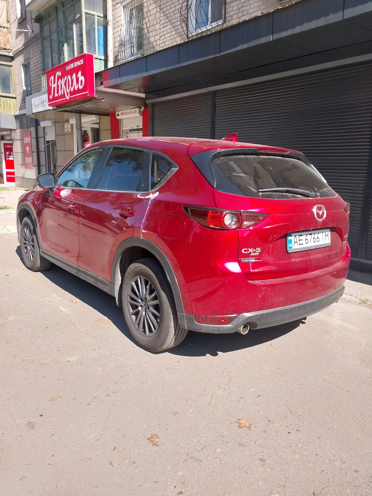 A red Mazda CX-5 is parked on a paved street in front of a building with a red sign that reads "Nikol." The car's license plate is AE 6766 IM. The building appears to be a commercial establishment, possibly a salon or shop, with large windows and roller shutters. The street is clean, with some fallen leaves on the ground. The sky is clear and the sun is shining, casting shadows on the street. The scene is typical of a city street in Dnipro, Ukraine.