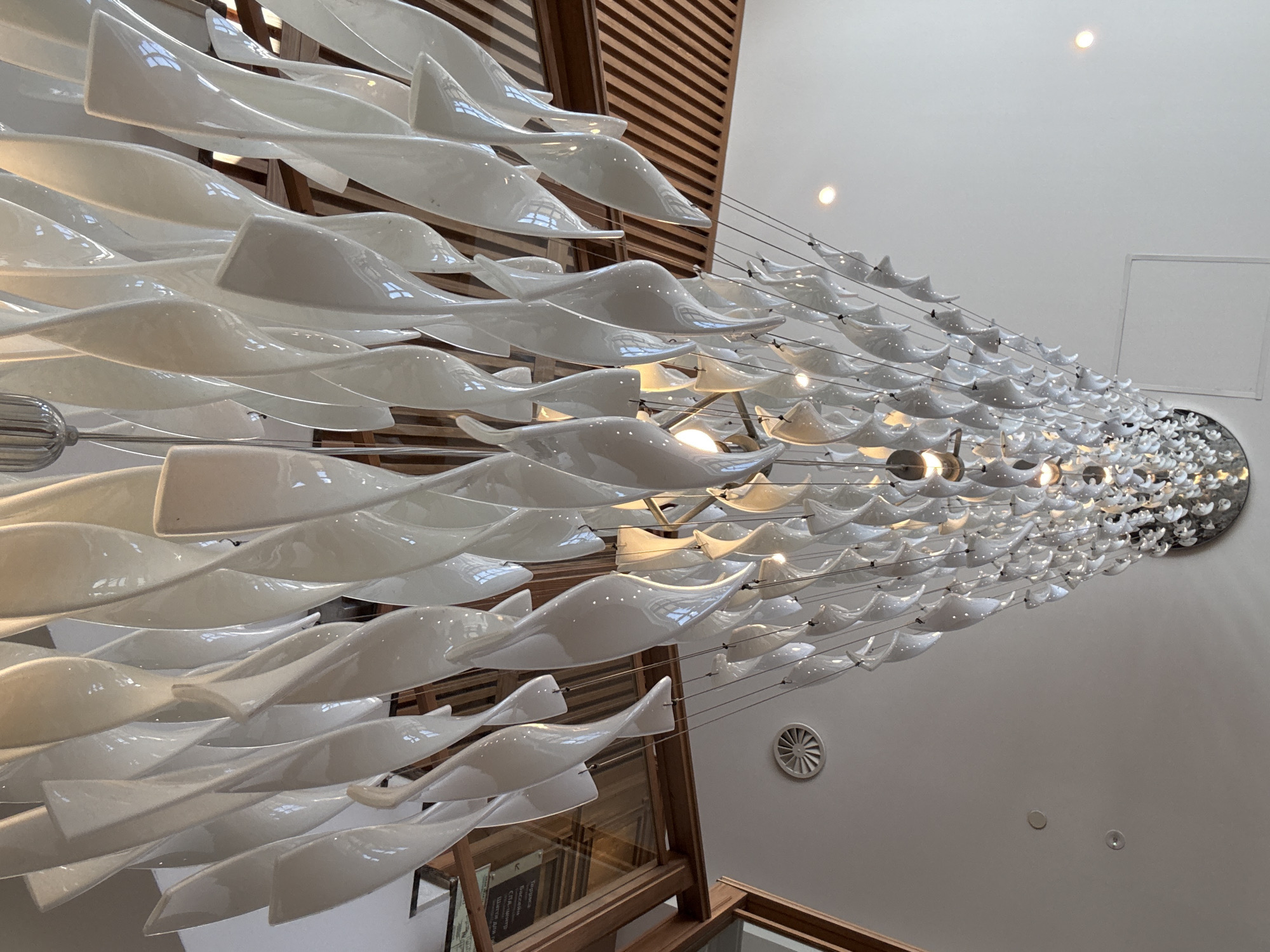 The image displays a large, elaborate chandelier or art installation dominating the upper portion of an indoor space. The fixture is composed of numerous individual elements, each a white, glossy, and curvilinear shape resembling an elongated wave, a twisted ribbon, or possibly a stylized fish. These sculptural components are suspended at varying heights by thin wires, forming a dense and tapering arrangement that extends diagonally across the frame. Integrated light sources within or behind these elements cast a warm glow, highlighting their reflective surfaces and creating intricate patterns of light and shadow.
In the background, a modern architectural setting is visible. On the left, a vertical structure made of warm-toned wooden slats rises, suggesting a wall or part of a ceiling design. The rest of the background is a plain, light-colored wall and ceiling. Several recessed spotlights are embedded in the ceiling, along with a circular ventilation grate mounted on a vertical surface. Towards the right, a square panel, possibly an access hatch, is visible on the ceiling.
No people are present in the image, focusing attention entirely on the artistic lighting fixture. The scene is brightly lit by both the fixture itself and ambient artificial lighting. On a lower level behind the wooden structure, a dark rectangular sign with unreadable text, appearing to be Cyrillic script, is partially visible. The overall impression is one of modern design and sophistication in a public or commercial building.