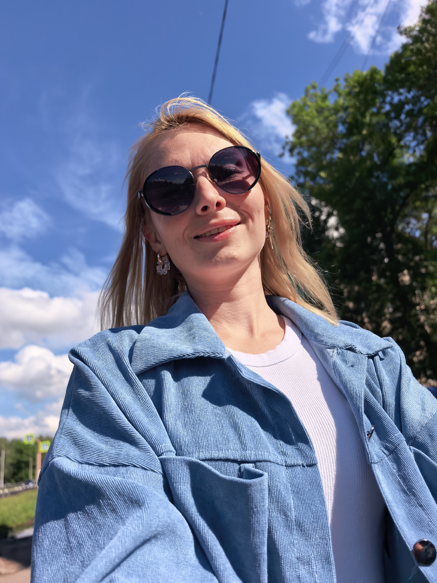 A low-angle, sunlit selfie portrait features a blonde-haired woman outdoors in Sterlitamak, Russia. She wears dark, round sunglasses reflecting the blue sky and green foliage. Her lips are slightly parted in a soft smile, and she has visible earrings: a multi-faceted clear earring on her left ear and a slender gold-toned drop earring on her right. Her hair shows faint light blue or green highlights towards the ends. She is dressed in an unbuttoned light blue corduroy jacket over a white ribbed top. The background consists of a bright blue sky with scattered white cumulus clouds, dense green tree foliage on the right, and overhead utility lines stretching across the upper frame. Distant, blurred traffic signals or signs and a road are visible on the far left.