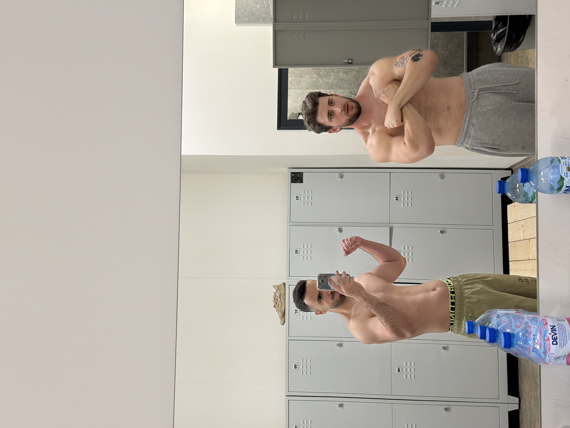 The image captures a mirror selfie taken in what appears to be a gym locker room. The scene is dominated by a large mirror on the left, reflecting a shirtless man wearing grey sweatpants. This man is flexing his biceps, with his arms crossed over his chest, and has a tattoo on his left upper arm. He has a beard and dark hair.
The man taking the photo is visible directly on the right side of the image. He is also shirtless, wearing olive green shorts with a visible elastic waistband featuring text. He holds a smartphone in his right hand to capture the reflection and is flexing his left bicep. He also has dark hair and a beard.
In the background, behind both men, are rows of grey metal lockers, some with numbers like "00", "22", and "23" printed on them. On top of the lockers near the man taking the selfie, a pair of light-colored sneakers are placed. Two plastic water bottles with blue caps, bearing the "DEVIN" brand name and "0.5L" capacity, are visible on a wooden surface in the lower right, with one partially seen in the reflection. A black trash bag is also visible in the upper right background of the reflection.
The setting is brightly lit, indicating an indoor environment. Both men appear to be showcasing their physique, likely after a workout. The presence of "DEVIN" water bottles suggests a location in Bulgaria, consistent with Plovdiv.