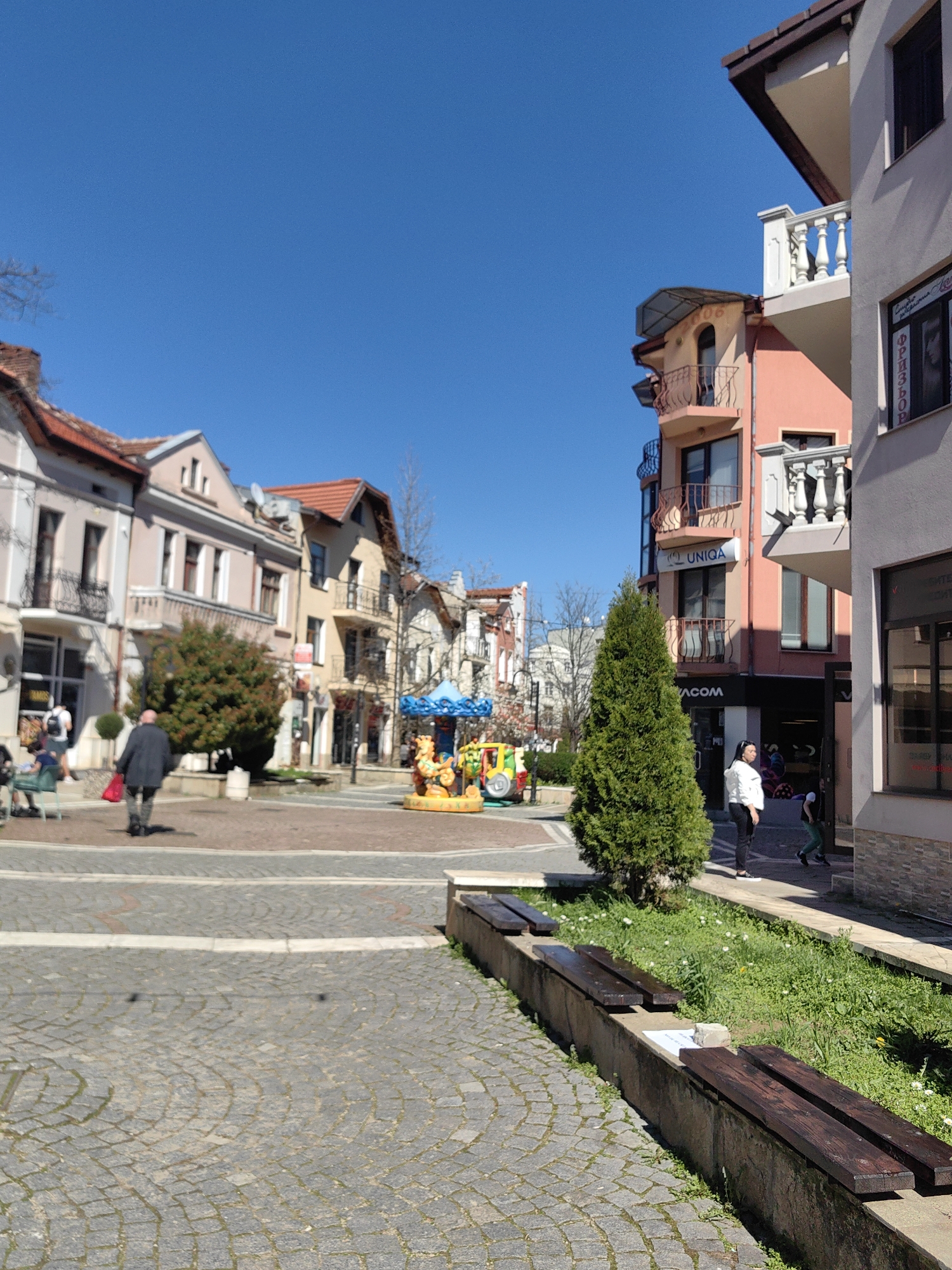 The image depicts a pedestrian square in Sevlievo, Bulgaria, with a small, colorful carousel in the center. The square is paved with gray cobblestones. Buildings with varying architectural styles line the square, including one with a UNIQA sign. Several people are visible, including individuals walking or standing near buildings. Benches made of concrete and wood are present in the foreground. The sky is clear and blue. Small trees and grass are planted near the benches.