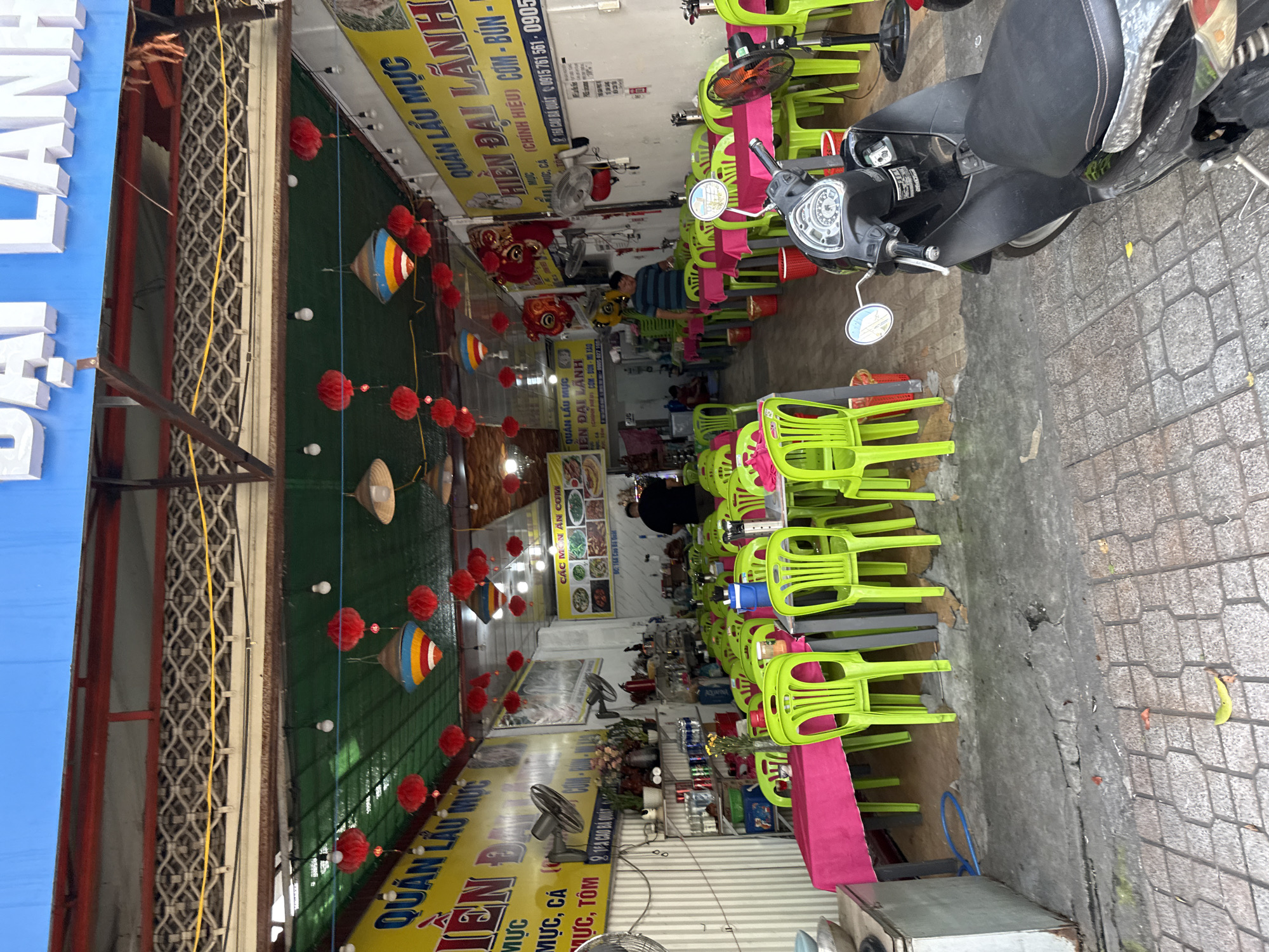 This is an exterior shot of a restaurant in Nha Trang, Vietnam, with the name "ĐẠI LÀNH" visible in large white letters on a blue awning. The restaurant appears to be an outdoor seating area with many bright green plastic chairs arranged around tables covered with pink tablecloths. The floor is a mix of concrete and patterned tiles, with some wear and tear visible.
Several red paper lanterns are hanging from the ceiling, along with some conical straw hats that also serve as decorative elements. There are fans mounted on the walls to help with ventilation. Signs in Vietnamese are displayed on the walls, including one that lists menu items and another with phone numbers.
A black scooter is parked in the foreground, partially obscuring the view of the restaurant's entrance. In the background, a few people are visible inside the restaurant, including a man standing near a table and another person in the distance. The overall impression is of a casual, local eatery.