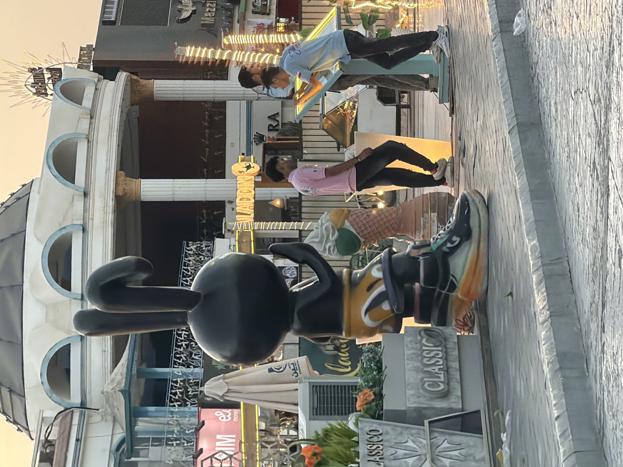 This image captures a street scene in Sharm El Sheikh, Egypt, likely in a commercial or entertainment district given the architecture and signage. In the foreground, a large, black cartoonish statue of a rabbit-like character wearing oversized sneakers and shorts stands prominently. To the right of the statue, three young men are gathered around a display panel, with two appearing to interact with it. One man in a pink shirt stands looking down, while two others in blue shirts are engaged with the display. Behind them, an ice cream cone statue is visible. Further in the background, a domed building with arched windows and classical columns forms part of the urban landscape, alongside various shopfronts and illuminated signage. The overall setting suggests a vibrant, possibly tourist-oriented, outdoor area with a relaxed, casual atmosphere.
