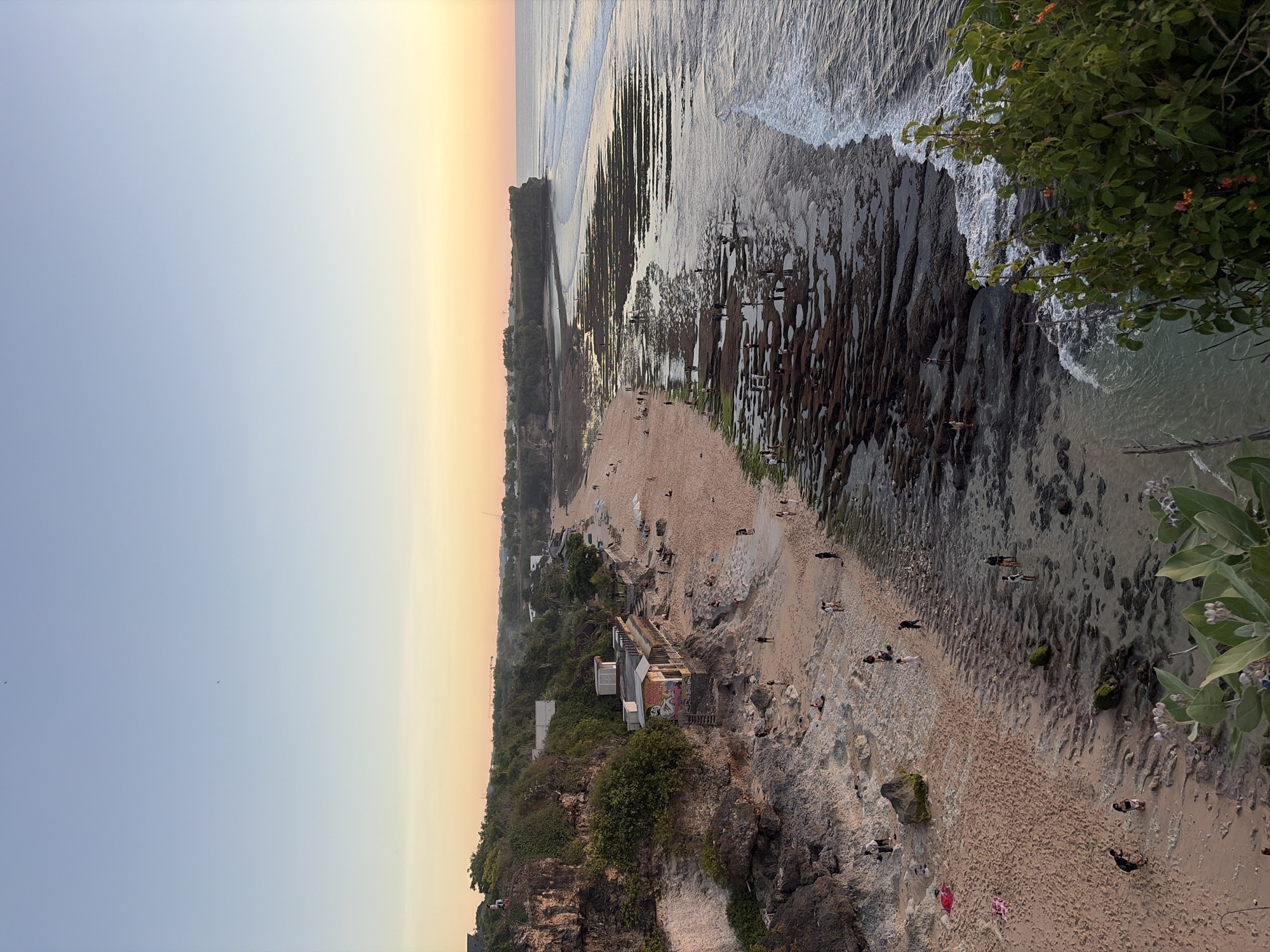 A high-angle, full shot captures a panoramic view of a tropical beach at sunset. The sky transitions from a clear, pale blue at the top to a soft orange-yellow glow near the horizon. The beach itself is a wide expanse of golden sand, dotted with rocks and patches of green moss near the water's edge. Numerous people are scattered across the sand and the exposed rocks, some walking, others appearing to be sunbathing or observing the scenery. The ocean is calm, with gentle waves breaking along the shore. Cliffs covered in lush green vegetation rise on the left side of the frame, with a few buildings nestled among the foliage. A vibrant green bush with small orange flowers is in the foreground on the right, partially obscuring the view of the rocky shoreline. The overall scene evokes a sense of tranquility and natural beauty.