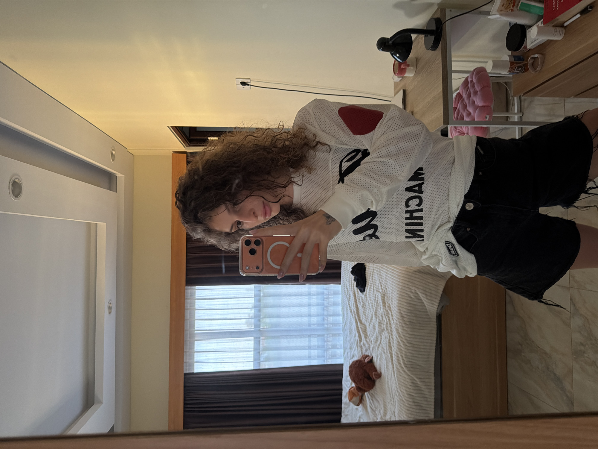 A young woman with curly brown hair stands in a bedroom, taking a mirror selfie. She is wearing a white long-sleeved mesh shirt with black text and a red heart patch on the sleeve, and black shorts. Her phone, with an orange case, is held up to take the photo. In the reflection, a bed with white bedding is visible, with a stuffed animal lying on it. Brown curtains hang over a window. To the right of the woman, a desk holds a black lamp, a mug, and other items. A pink stool is beneath the desk. The room appears to be a hotel room or a bedroom in a residential building. The floor is tiled. The image is likely taken in North Kuta, Indonesia, as indicated by the location tag associated with the image.