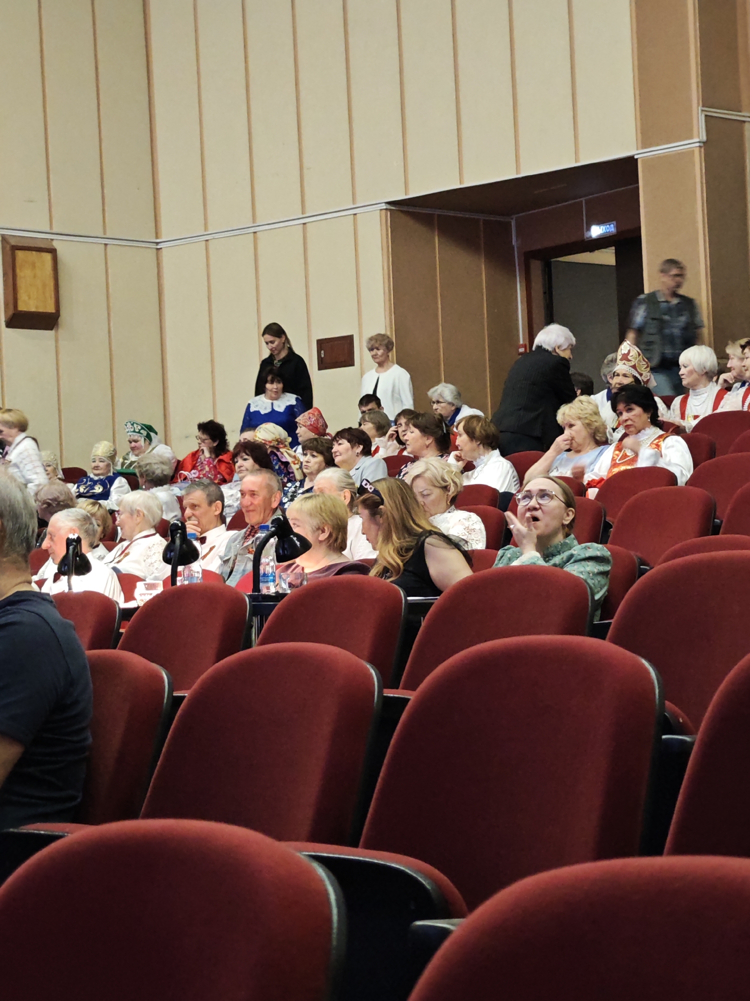 The image shows an audience in a theater or auditorium, likely in Марковский, Russia. The audience members are seated in red, cushioned chairs, and are mostly older adults, including some women wearing traditional Russian clothing such as embroidered shirts and headscarves. Many are seated on a raised tier. A man in a casual shirt is seen walking through a doorway at the back of the room, beneath an exit sign that is written in Cyrillic script.