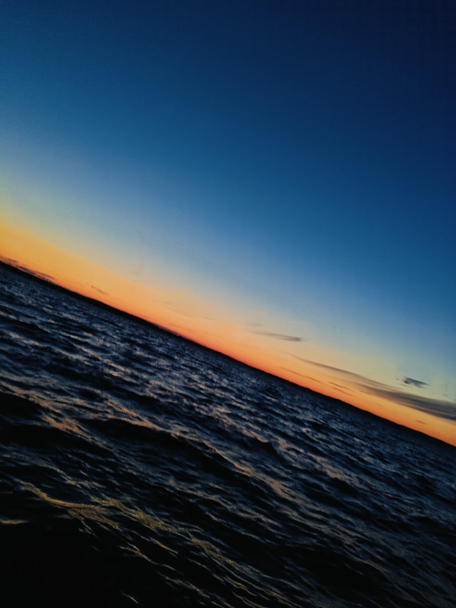 The image is a tilted, low-angle shot of a body of water under a gradient sky. The sky transitions from a deep blue at the top to a pale orange and yellow band near the horizon, suggesting dusk or dawn. The water is dark, with visible ripples and reflections of the sky's colors. The diagonal orientation of the scene is a prominent feature. The location is identified as Chaykovsky, Russia. No people, specific objects, or actions are discernible in the image.