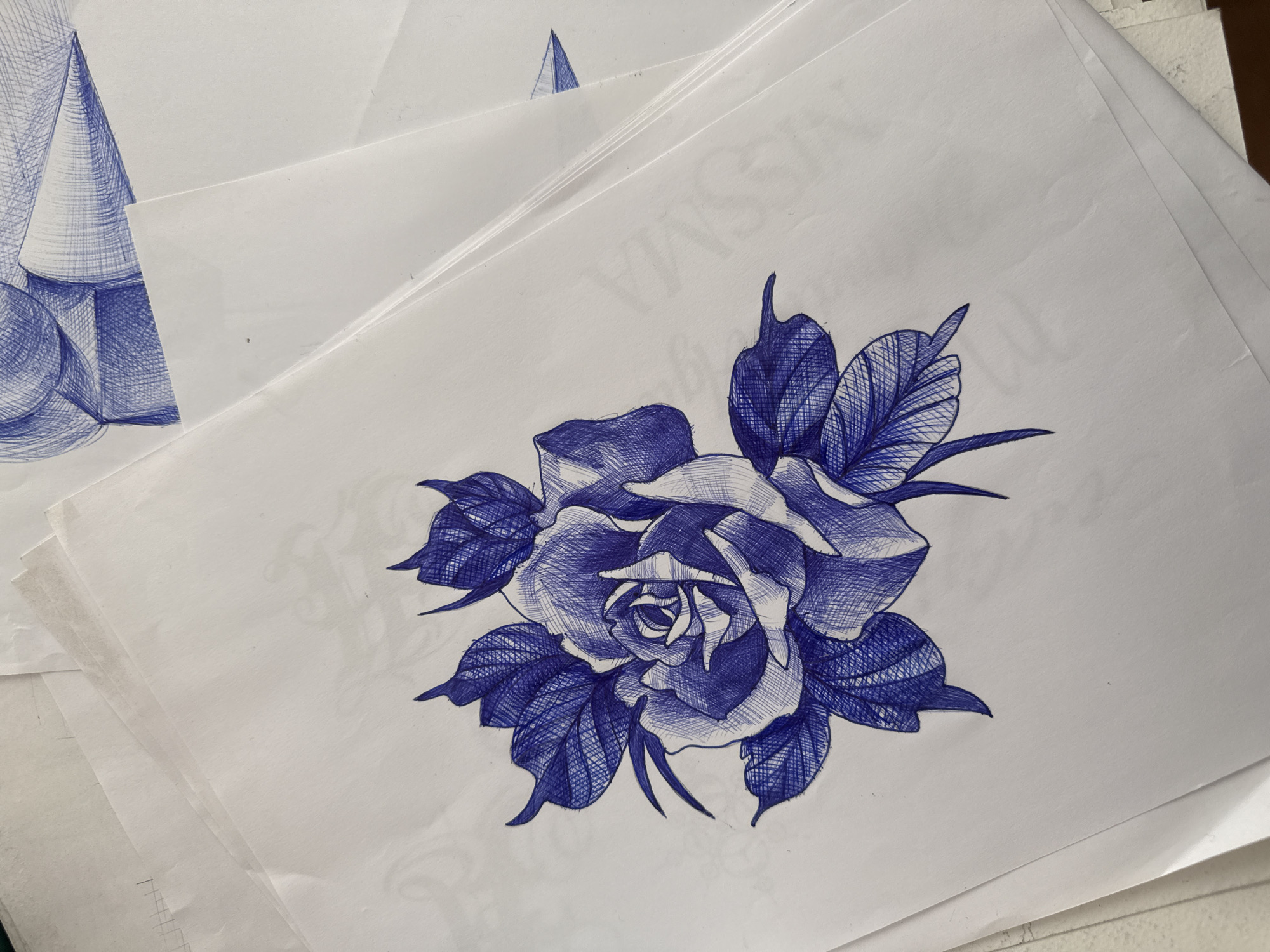 The image depicts a stack of white papers with blue ink drawings on top. The most prominent drawing is a rose with leaves, rendered in a cross-hatching style. Another paper shows geometric forms, a cone and a sphere, also in blue ink. A small blue triangle is also seen on one of the papers. The scene appears to be a workspace with sketches. Location information is not evident from the image.