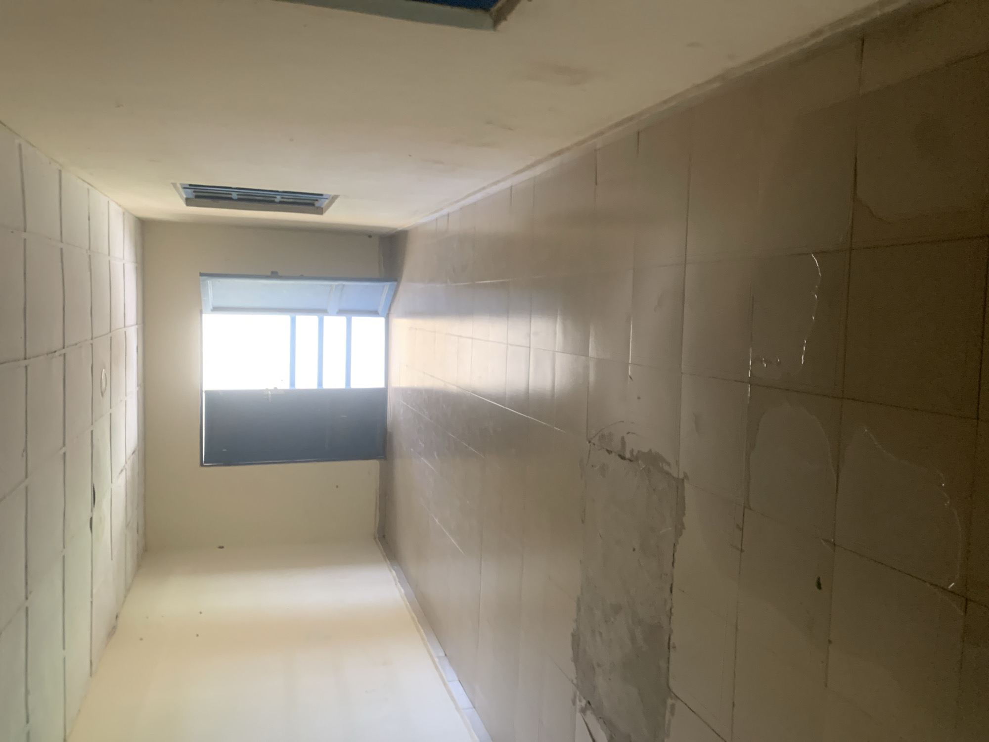 This media file depicts an interior view of what appears to be an unfinished or under-renovation room. The scene is characterized by bare walls and a tiled surface on the right side of the frame, which shows some signs of wear and tear, including patches of plaster or sealant. On the left, the wall is covered in what looks like white paneling or a similar material, also with visible lines or seams.
A prominent feature is a window with white blinds, letting in bright natural light, suggesting it is daytime. The lighting is diffused and somewhat harsh, typical of daylight entering an interior space. There is a ventilation grate installed in the ceiling, indicating it is a functional room. The floor appears to be a smooth, light-colored surface.
The room has a utilitarian feel, lacking any furniture or decorative elements. The overall impression is one of a space in transition, possibly a hallway or a room within a building undergoing construction or refurbishment. There are no people visible in the image, and no specific activity is taking place. The setting appears to be a standard indoor environment, with no strong indicators of weather conditions outside. The location context of Jalingo, Nigeria, does not provide specific visual cues within the image itself. No text is visible in the media file.