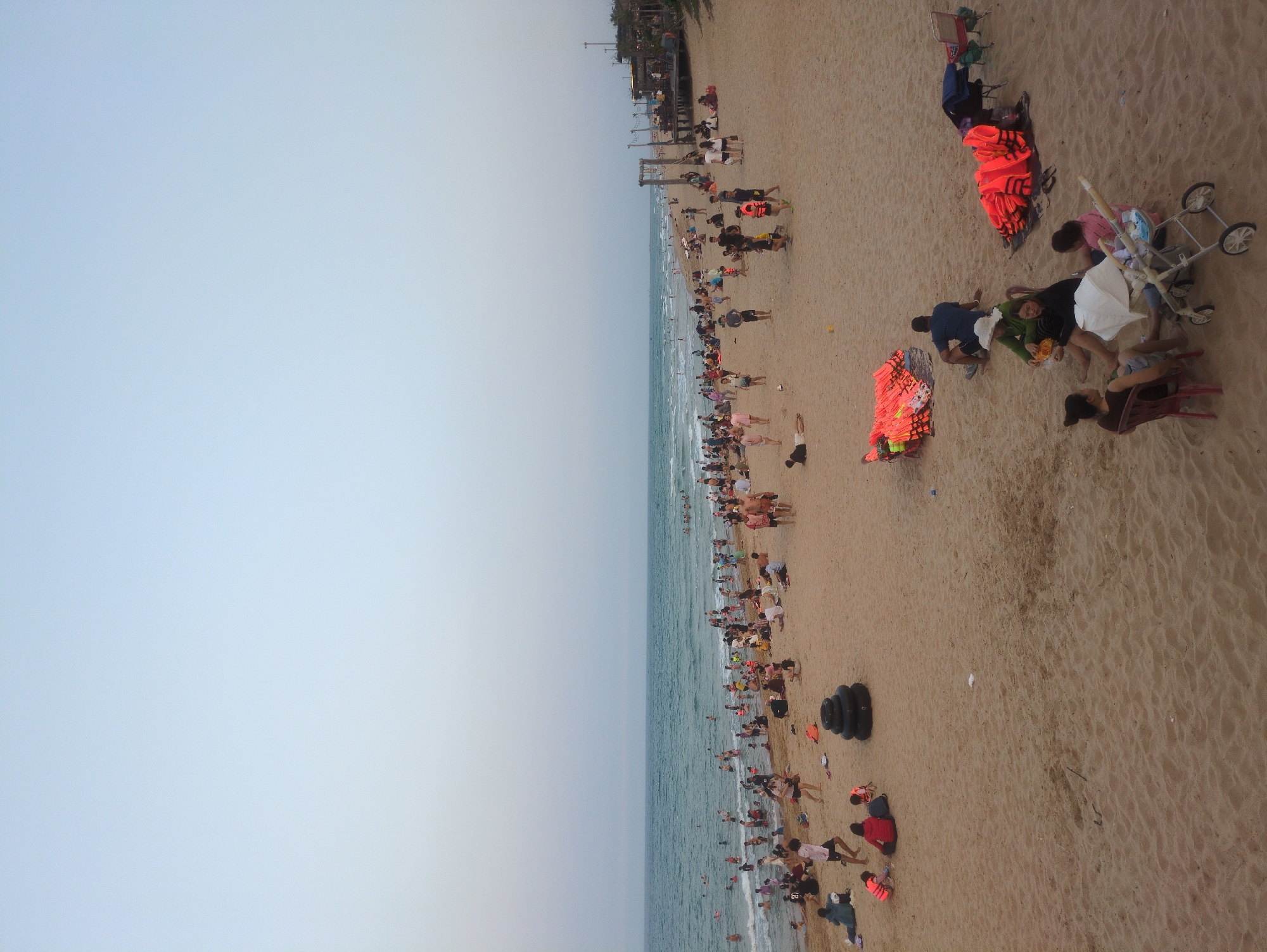 The image shows a crowded beach in Binh Kien, Vietnam. A large expanse of light blue sky is visible on the left side of the image. The ocean meets the sandy beach, where many people are swimming, wading, and sunbathing. There are stacks of bright orange life vests on the beach, along with several black inner tubes. Groups of people are gathered on the sand near umbrellas, and at least one stroller is present. Trees and structures are visible along the top edge of the frame.