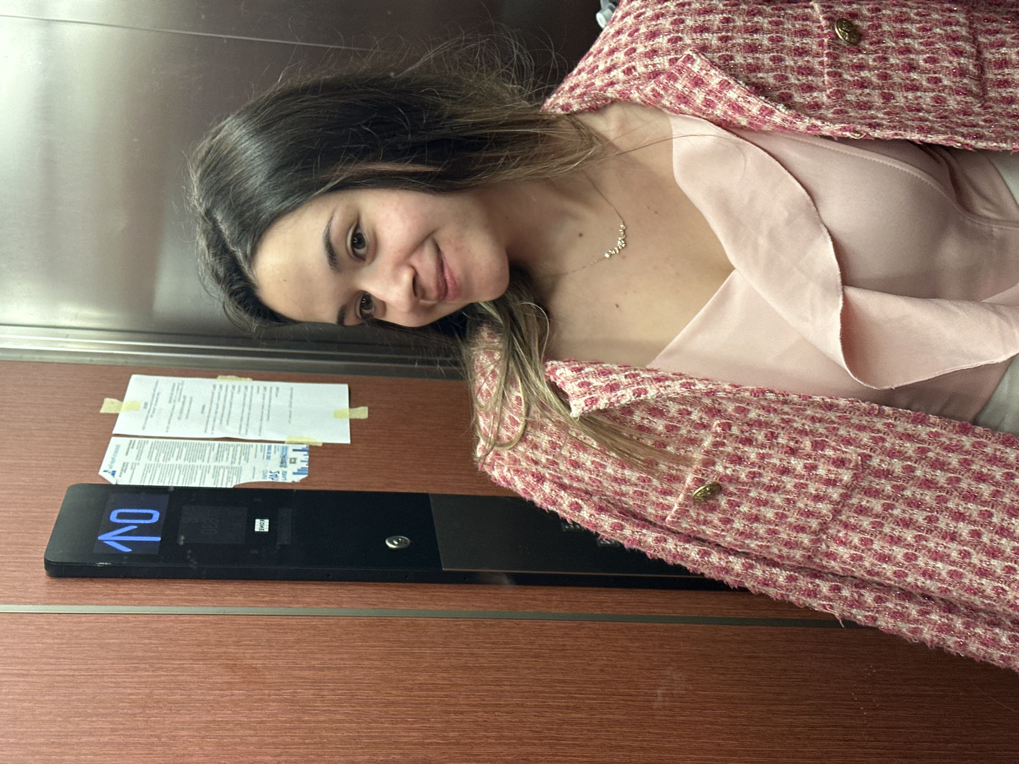 Inside an elevator in Sofia, Bulgaria, a young adult female with long, dark brown hair and a gentle smile poses slightly away from the camera. She is dressed in a light pink ruffled camisole or top, covered by an open, textured pink and white tweed-style jacket with gold-colored buttons on its pockets. A delicate gold-colored necklace rests on her chest. The elevator's interior features reddish-brown wood paneling with vertical grain on the side walls, separated by a metallic strip, and a reflective metal back wall. A black control panel on the right displays a bright blue upward arrow and the number "10", indicating the elevator's current or destination floor. Distorted text below the display reveals "KONE". Several notices, primarily written in Bulgarian (e.g., "СУТРИН", "ПОНЕДЕЛНИК", "ПЕТЪК"), are taped to the right wood panel with yellow adhesive.