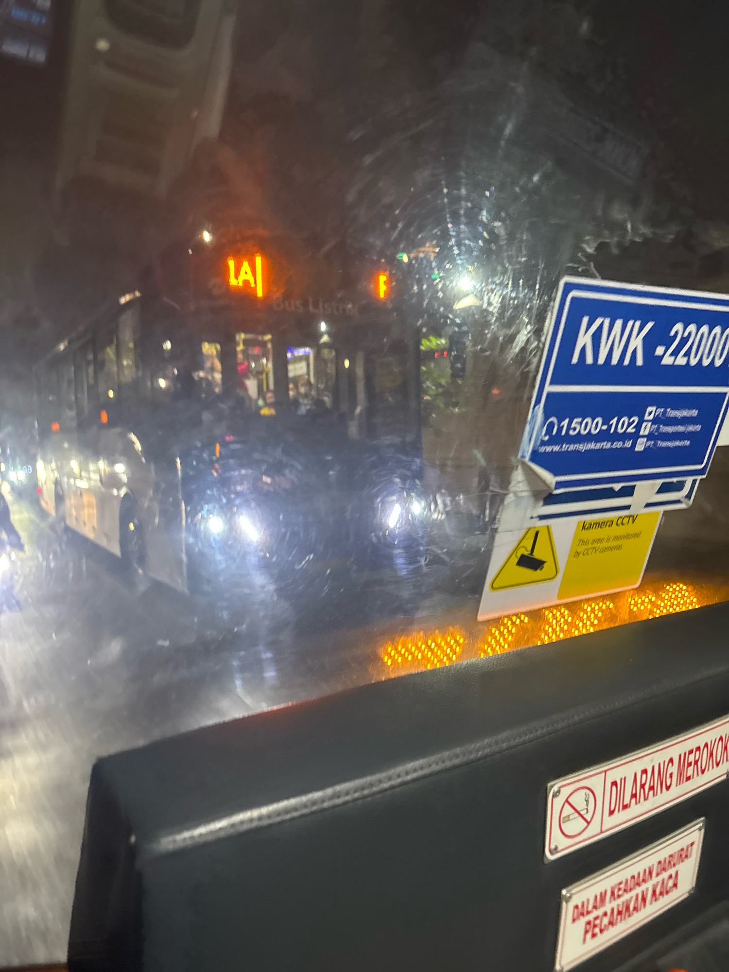 The image captures a scene from inside a bus in North Jakarta, Indonesia, at night. The bus appears to be illuminated by the headlights of other vehicles and streetlights, reflecting off the wet windshield. On the windshield, several signs are visible. A blue sign on the right reads "KWK -22000" and includes contact information for PT Transjakarta, a public transport company in Jakarta. Below this, a yellow sign with a black border features a graphic of a CCTV camera and the text "kamera CCTV" and "This area is monitored by CCTV cameras."
Inside the bus, passengers are visible through the windows, appearing as indistinct figures in the dim light. The bus itself has illuminated signage, including one displaying "LA" in orange, and other text that is difficult to decipher due to the image quality and reflections. Towards the foreground, a black padded surface is visible with two signs attached. One sign, with a red border, depicts a crossed-out cigarette and reads "DILARANG MEROKOK," meaning "No Smoking." Below this, another sign with a red border reads "DALAM KEADAAN DARURAT PECAHKAN KACA," which translates to "In Case of Emergency Break Glass." The overall atmosphere suggests a busy urban environment during nighttime transit.