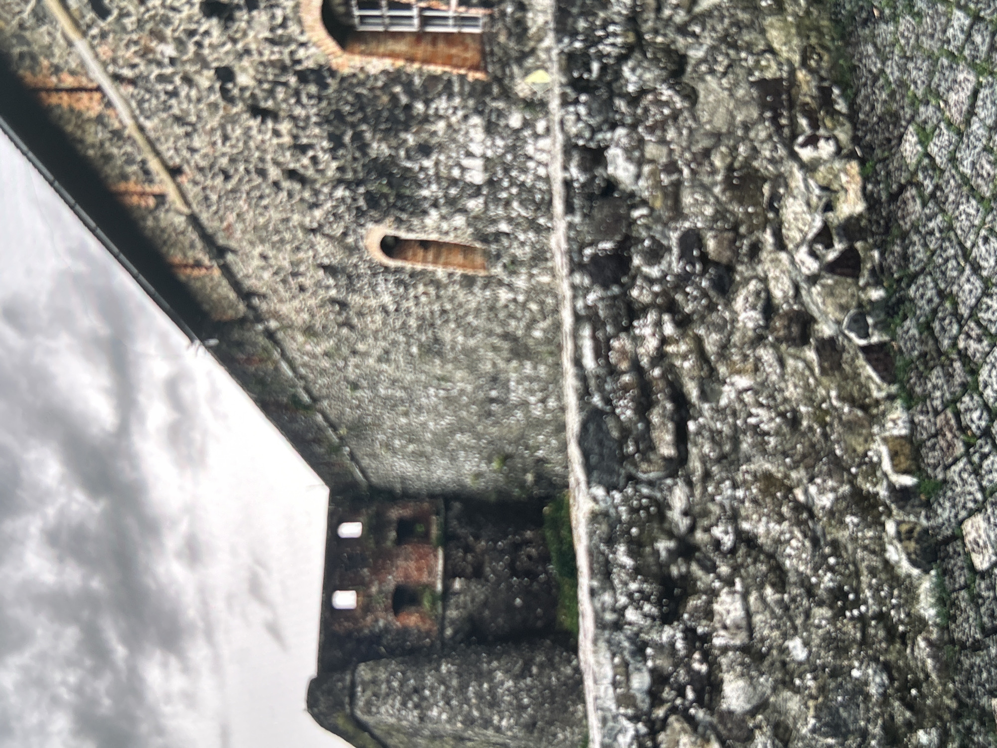 The image depicts a section of a historic stone fortification, likely part of Uzhhorod Castle in Uzhhorod, Ukraine. The fortification is constructed from large, irregular grey stones, giving it a rugged and ancient appearance. In the background, a portion of a building with red brick accents is visible, featuring several small, rectangular windows and arched openings. The sky above is overcast with dramatic, dark grey clouds, suggesting a somber or stormy atmosphere. In the foreground, a textured stone wall runs horizontally across the frame, with a cobbled pathway partially visible at the bottom, interspersed with patches of green moss or grass. The overall scene is devoid of any people or animals, focusing solely on the architectural elements and the imposing nature of the fortification under a heavy sky.