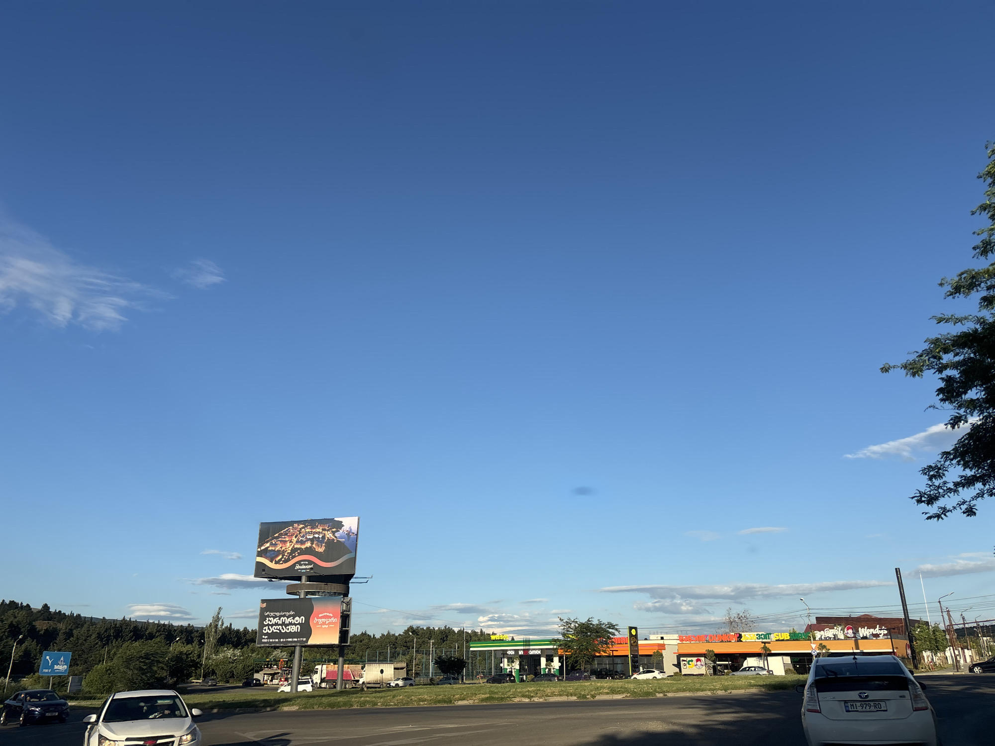 This media file captures a daytime scene on a road in Tbilisi, Georgia. The sky is a vibrant blue with a few wispy clouds, suggesting pleasant weather. The foreground shows a paved road with several cars, including a white Chevrolet and a white Toyota Prius. In the middle ground, a billboard displays an image of a city at night, with text in Georgian. Next to it, another billboard features text and imagery, also in Georgian. Further back, a cluster of businesses lines the road, including a gas station with green and white branding, a building with signage for "Wendy's" and "Subway," and other commercial establishments. Trees and a wooded area are visible in the background, adding to the suburban or roadside commercial setting. The overall impression is a typical urban or peri-urban environment during daylight hours, with everyday traffic and commercial activity.