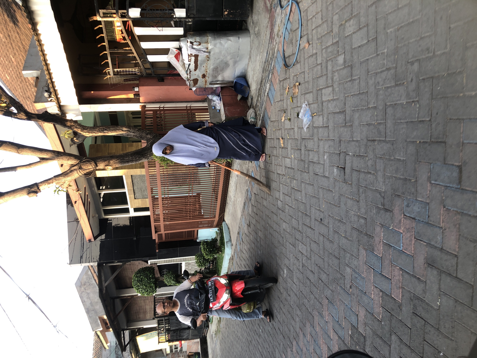 In the city of Kalisampurno, Indonesia, a man rides a red motorcycle down a paved street. He wears a grey t-shirt with a graphic print and blue jeans, with a pair of black sandals on his feet. Behind him, a woman in a long blue dress and a light blue hijab sweeps the street with a broom. Her feet are adorned with pink sandals. The street is paved with interlocking grey bricks. The background features houses with terracotta tile roofs and brown metal fences. To the left, there are some green bushes and a tree. To the right, there's a large metal bin filled with refuse, and a blue hose is coiled on the ground. The scene appears to be in the late afternoon, with soft sunlight casting shadows.