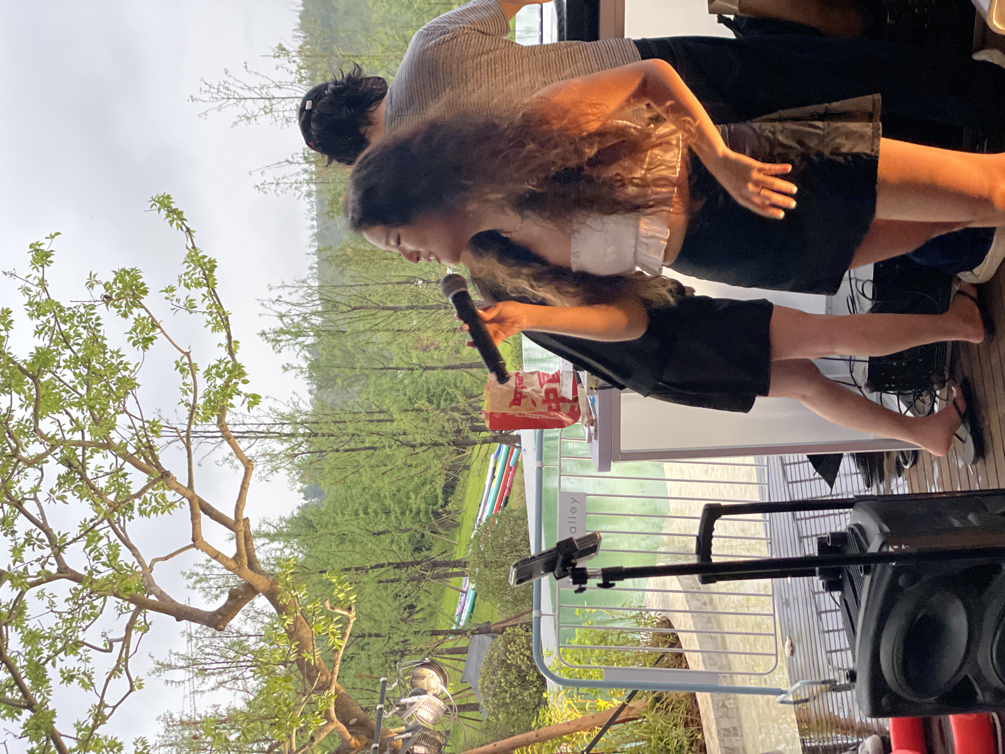 In Liangjiang, China, a young woman with long, curly brown hair is captured mid-song, holding a microphone and singing into it. She is wearing a white ruffled crop top and a black skirt, with a ring on her right hand. Behind her, another person, partially obscured, stands with their back to the camera, wearing a striped shirt. They appear to be near a white portable sound booth or equipment setup. To the left, a mobile phone is mounted on a stand, and a black speaker sits on the wet wooden deck. The background reveals lush greenery with numerous trees and a body of water, possibly a canal or pond, with several colorful paddleboards or kayaks arranged on a grassy slope. Overhead, branches of a tree with green leaves frame the scene against a cloudy sky. The overall impression is an outdoor performance or event in a natural setting.