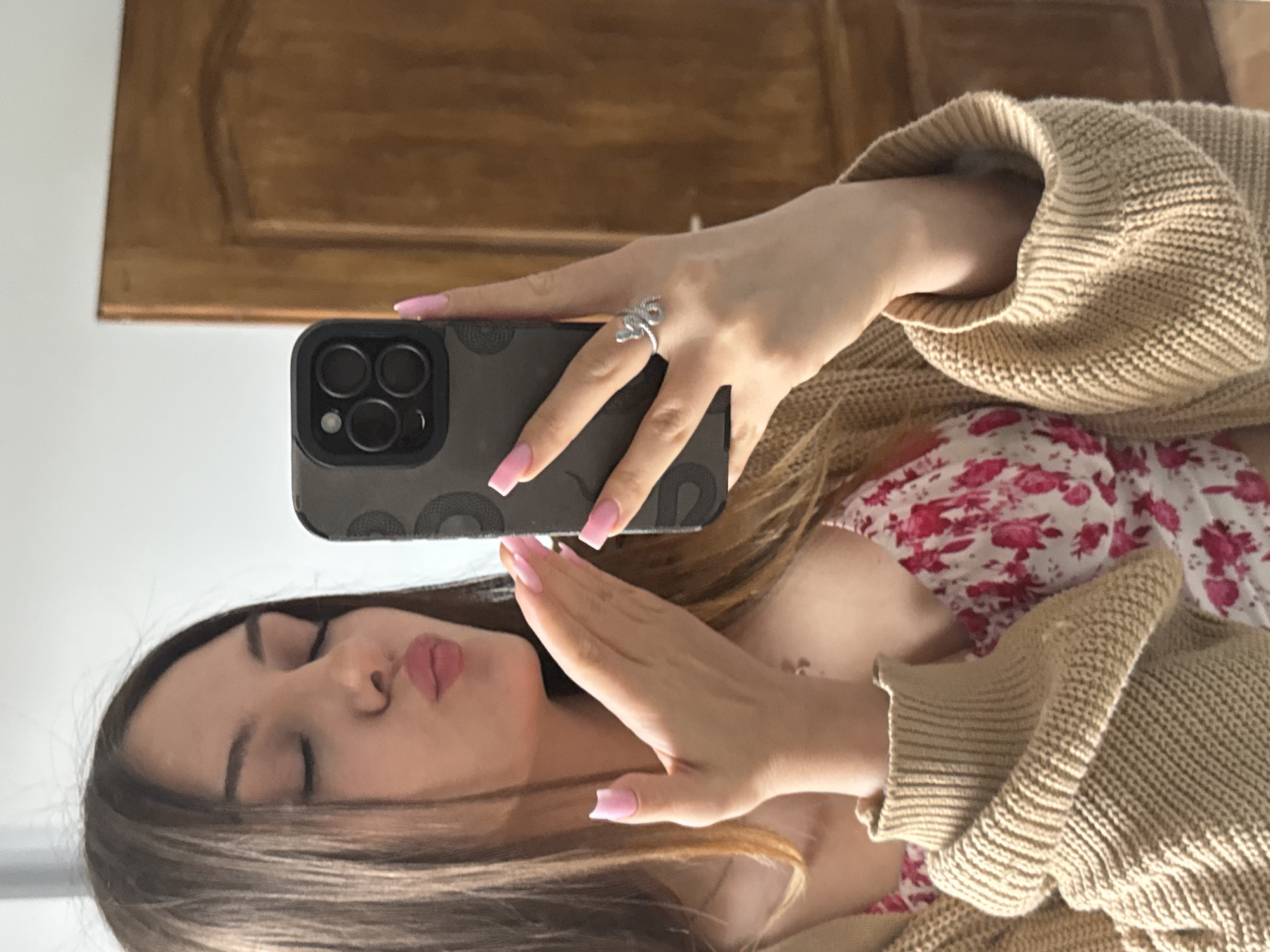 A young woman with long brown hair is taking a selfie with her phone. She is wearing a beige knitted sweater and a white floral dress. She has pink nail polish and a silver ring on her finger. The background appears to be a wooden door. The location is unknown and there is no way to ascertain whether this image was taken in Sofia, Bulgaria.