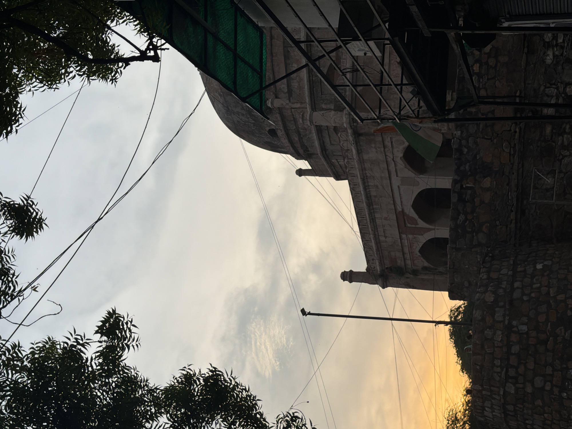 The image depicts a historic domed structure in Delhi, India, likely a tomb or monument, under a cloudy sky at sunset. The architecture features arches and ornate stonework. In the foreground, a rough-hewn stone wall forms a lower level. Electrical wires are visible strung across the sky, and some tree branches frame the scene from the top. A metal structure with a green canopy and railings is partially visible on the right, and an Indian flag is attached to a pole. The sunset casts a warm glow on the sky in the background.